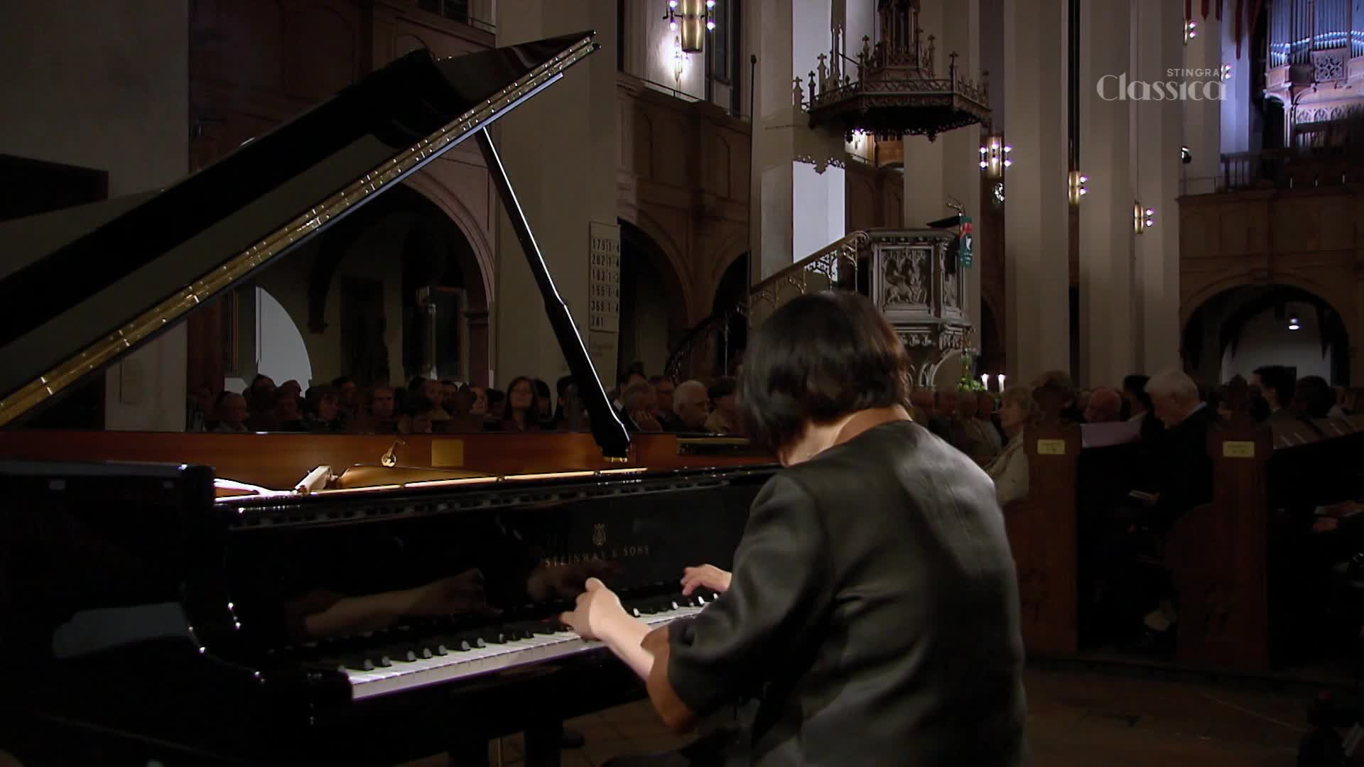 A pianist's hands move across the keys of a grand piano, filling the hushed church with music.  An audience watches intently from the pews, bathed in the warm glow of the sanctuary.
