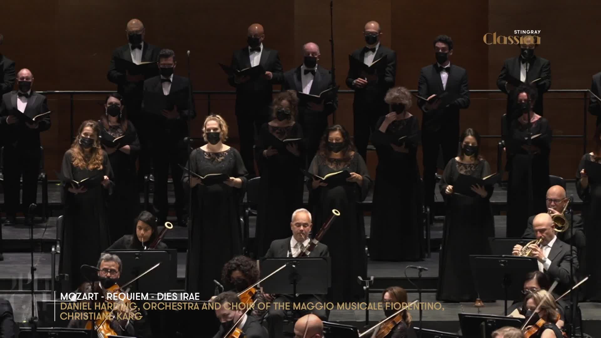 A choir in black gowns stands on risers, holding sheet music. Below them, an orchestra is in full swing, with musicians playing various instruments.