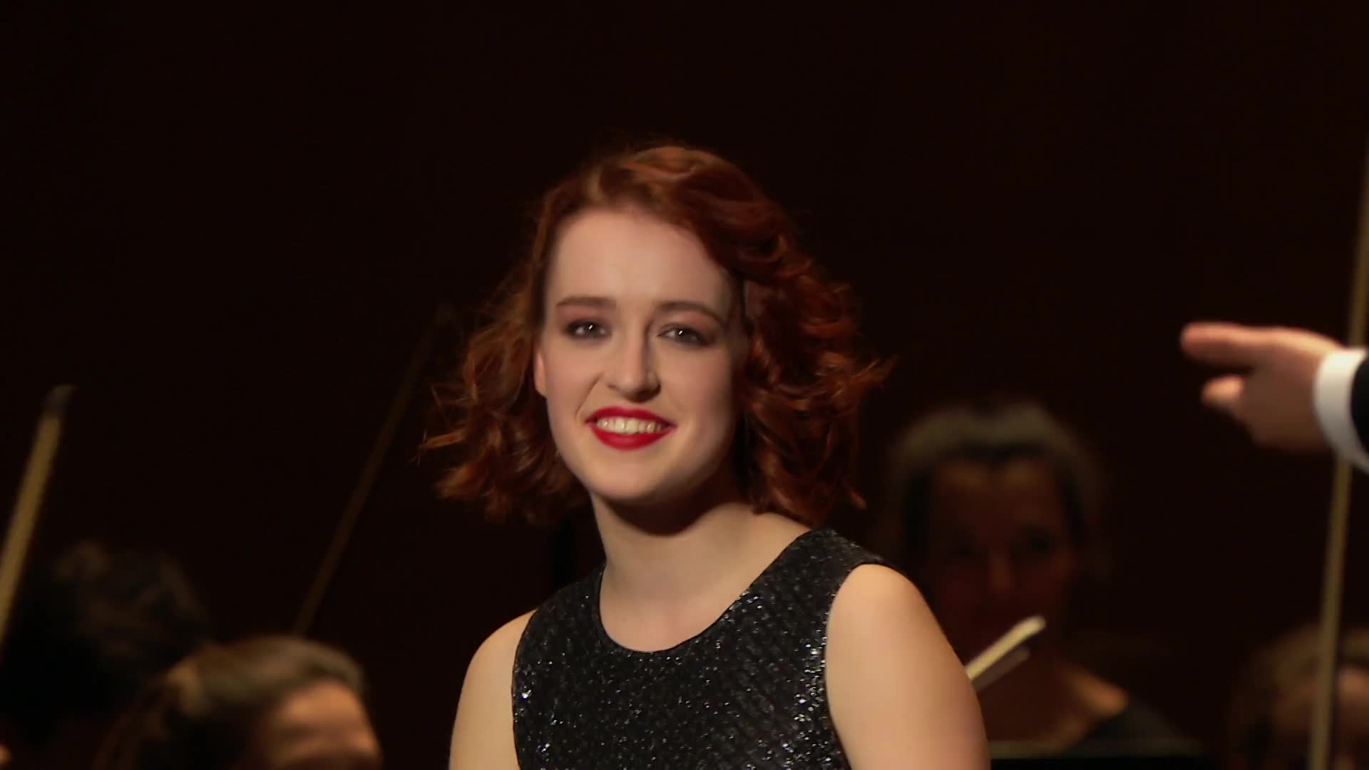 A young woman with bright red hair smiles, her sequined dress catching the light. Behind her, a conductor gestures with his baton, leading an orchestra through their performance for Stingray Classica.
