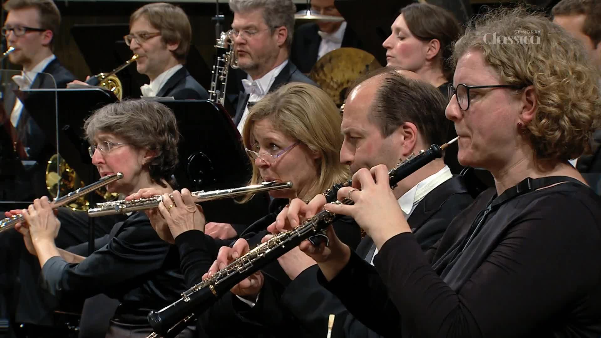 The flutist in front, a woman with short, curly hair, plays a silver flute. Beside her, a man with glasses holds an oboe to his lips.