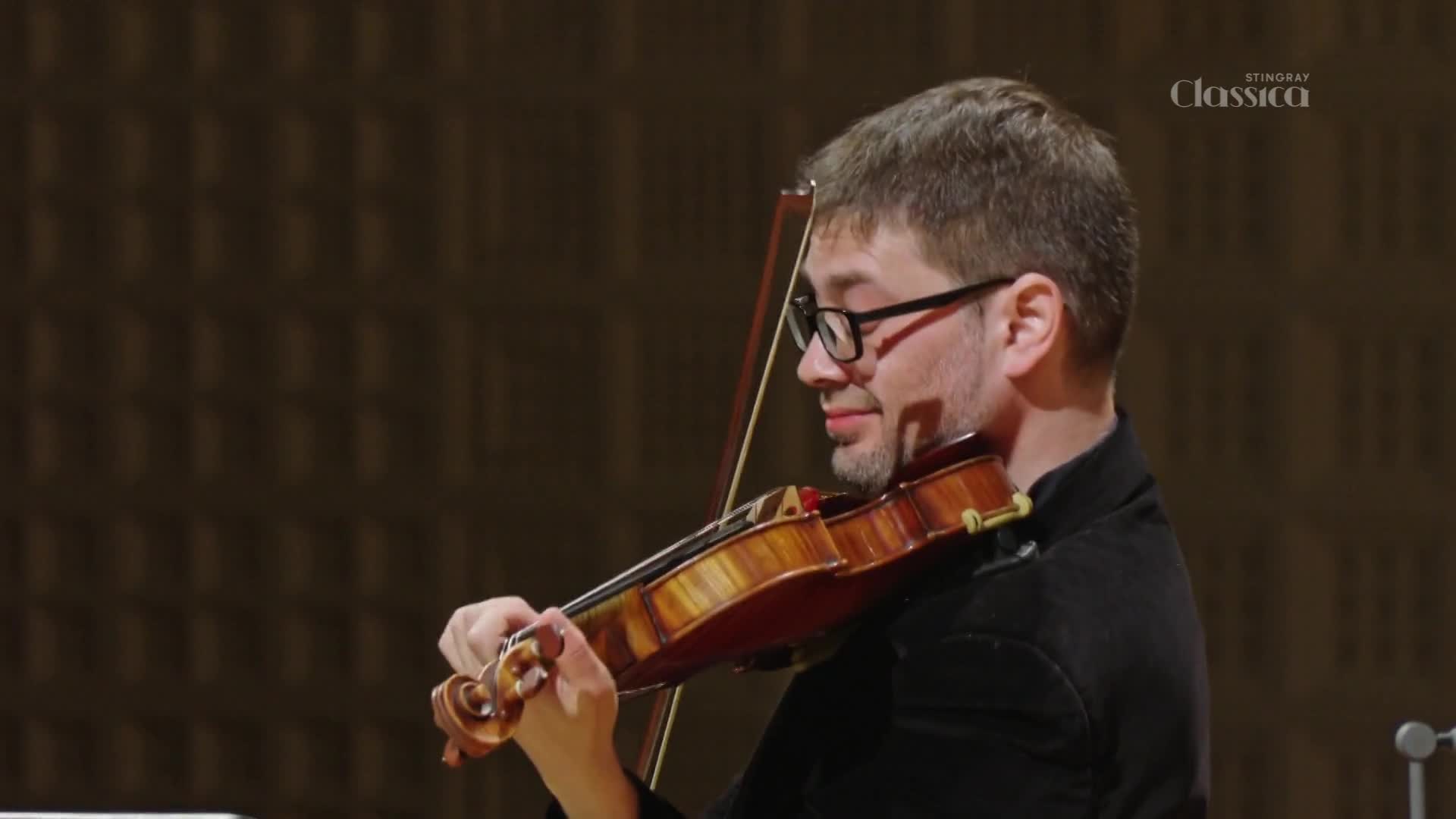 A man in glasses draws his bow across a violin, his face intent. The rich wood of the instrument gleams under the stage lights.