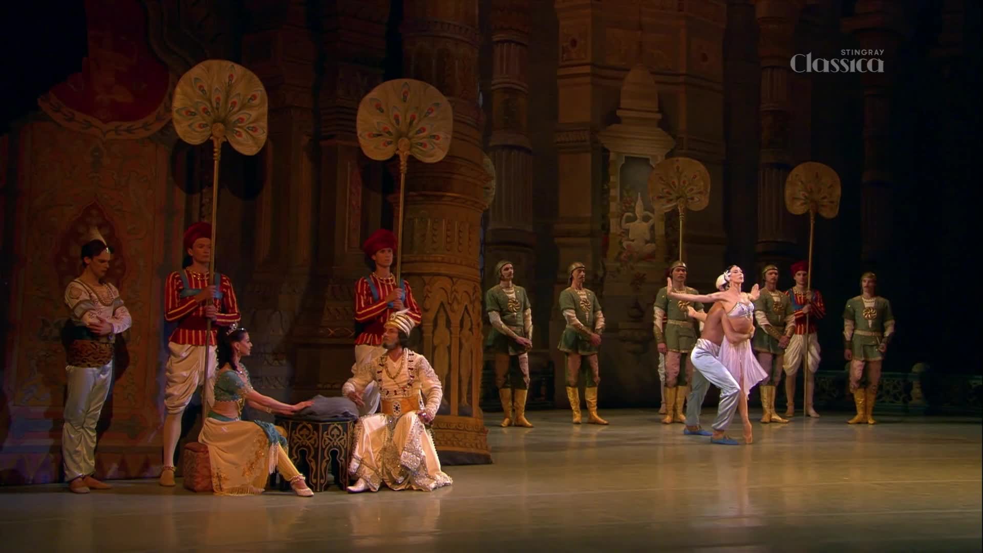A male dancer lifts his female partner, her pink tutu flaring as they move across the stage. Guards in green tunics and red-capped attendants stand at attention, holding large, ornate fans.