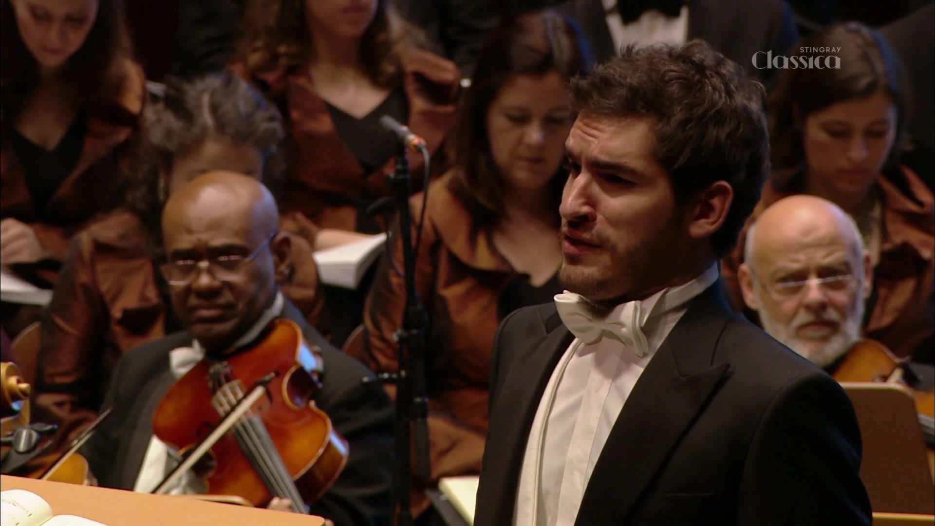 A tenor sings with his chest out, his white bow tie stark against his black tuxedo. Beside him, a violinist with glasses draws his bow across the strings, his instrument gleaming.