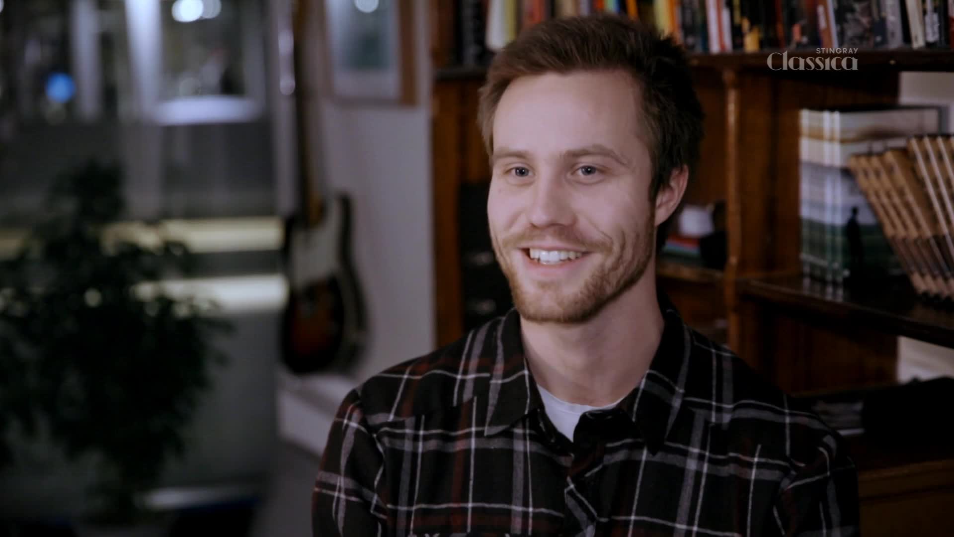 A young man with a beard grins, his eyes crinkling at the corners. Behind him, bookshelves overflow with titles, and a guitar hangs on the wall. A young man with a beard grins, his eyes crinkling at the corners. Behind him, bookshelves overflow with titles, and a guitar hangs on the wall.