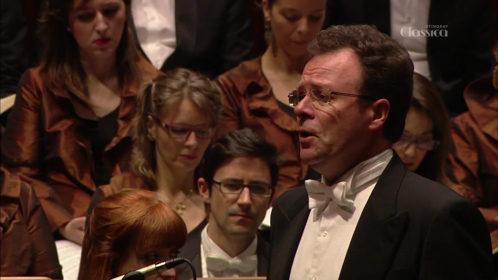 A man in a tuxedo with a white bow tie sings, his mouth open as he performs. Behind him, a choir in matching brown jackets looks on, some with their heads bowed. A man in a tuxedo with a white bow tie sings, his mouth open as he performs. Behind him, a choir in matching brown jackets looks on, some with their heads bowed.