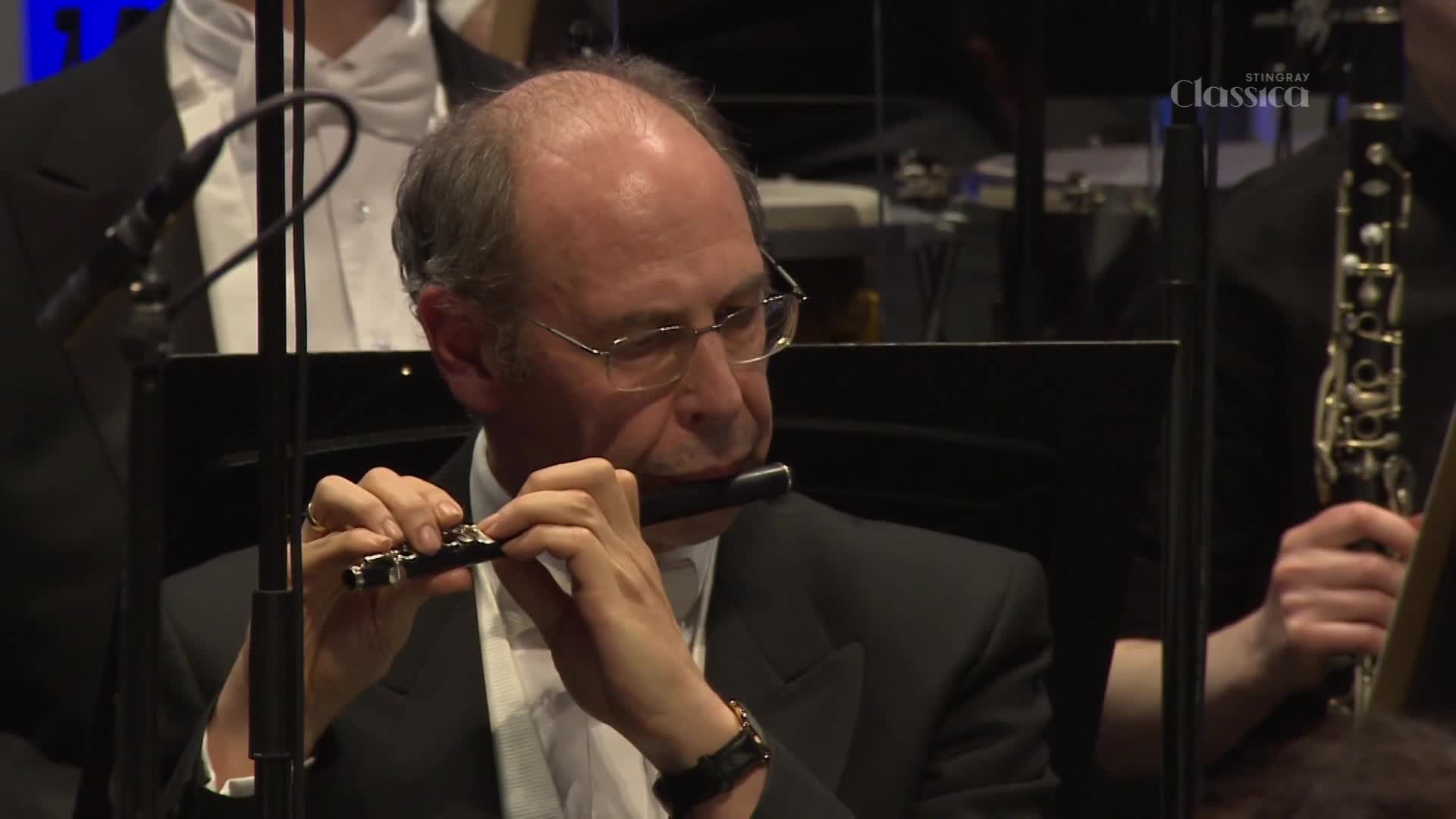 A man in a tuxedo plays a dark flute, his fingers moving nimbly over the keys. Behind him, other musicians in formal wear are visible, their instruments catching the light. A man in a tuxedo plays a dark flute, his fingers moving nimbly over the keys. Behind him, other musicians in formal wear are visible, their instruments catching the light.