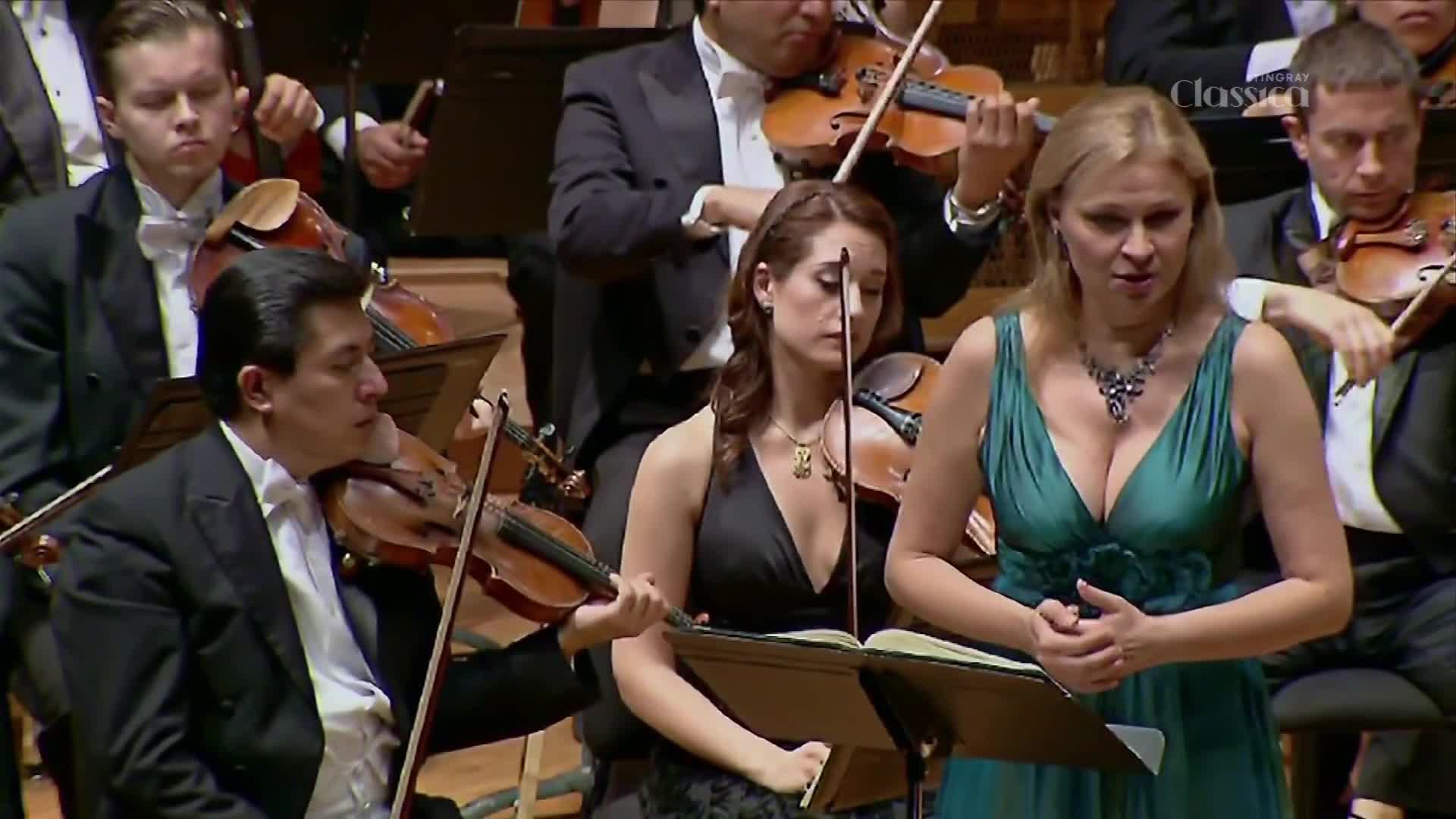 A soprano in a teal gown sings with her hands clasped, while a violinist in a black dress plays beside her. The orchestra behind them, dressed in formal wear, continues their performance. A soprano in a teal gown sings with her hands clasped, while a violinist in a black dress plays beside her. The orchestra behind them, dressed in formal wear, continues their performance.