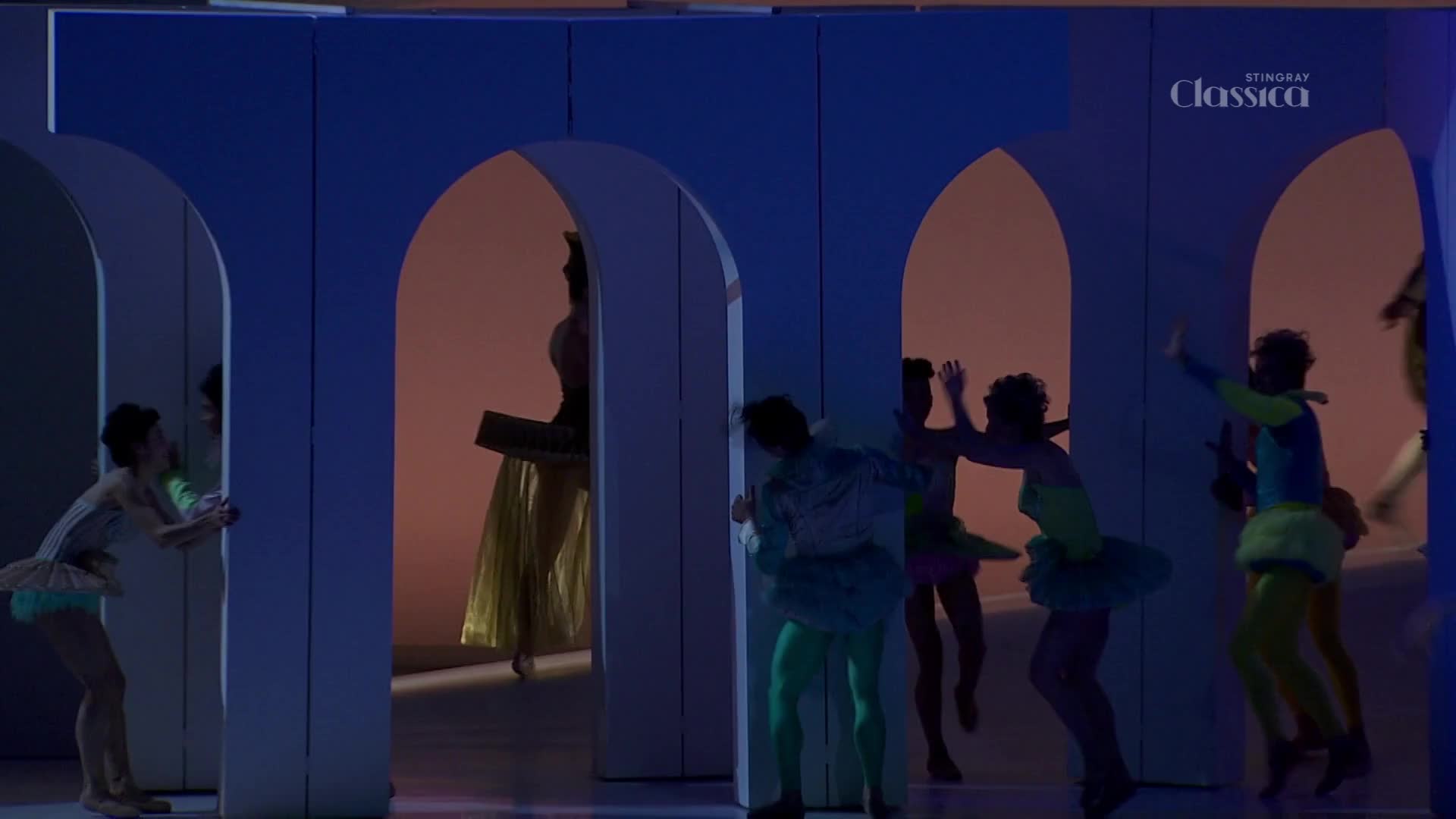 Dancers in bright, varied costumes move through arched openings on a stage. One dancer, silhouetted in gold, walks through a distant archway. Dancers in bright, varied costumes move through arched openings on a stage. One dancer, silhouetted in gold, walks through a distant archway.