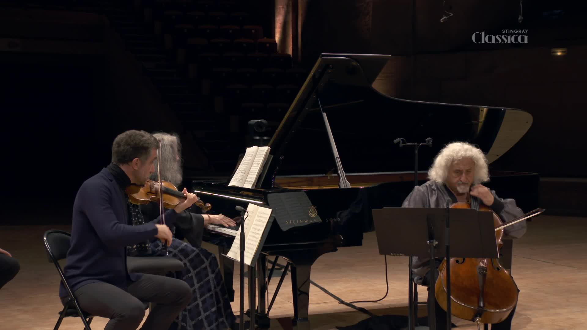 A violinist, seated to the left, plays with focused intensity, his bow gliding across the strings. Beside him, a pianist's hands move over the keys of a grand piano, their fingers a blur of motion. Across the stage, a cellist with a shock of white hair draws his bow across his instrument, his eyes closed in concentration. The Stingray Classica logo is visible in the upper right corner. A violinist, seated to the left, plays with focused intensity, his bow gliding across the strings. Beside him, a pianist's hands move over the keys of a grand piano, their fingers a blur of motion. Across the stage, a cellist with a shock of white hair draws his bow across his instrument, his eyes closed in concentration. The Stingray Classica logo is visible in the upper right corner.