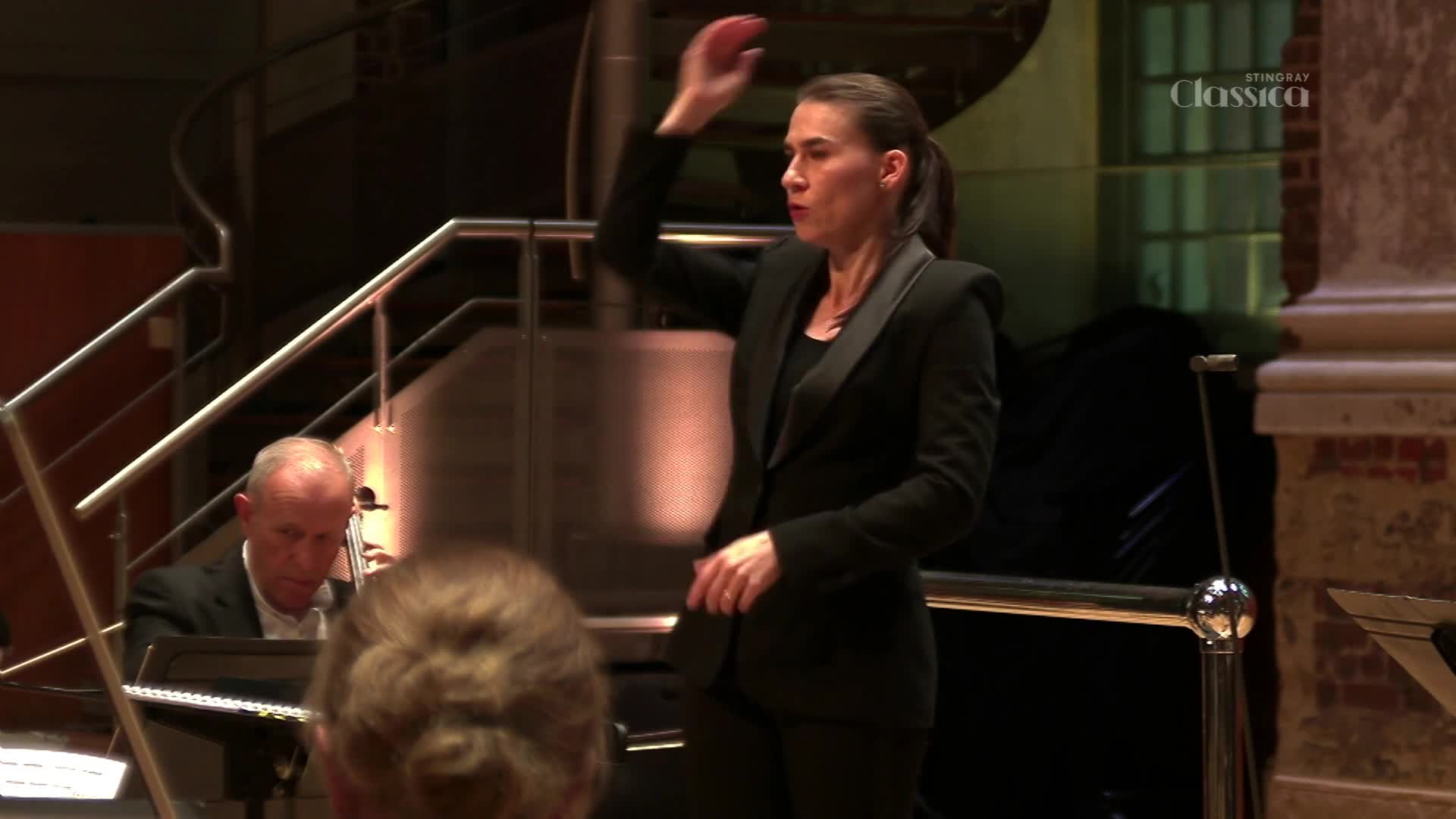 The conductor raises her arm with a sharp gesture, leading an orchestra in what appears to be a performance for Stingray Classica. A pianist, focused on his music, sits nearby. The conductor raises her arm with a sharp gesture, leading an orchestra in what appears to be a performance for Stingray Classica. A pianist, focused on his music, sits nearby.