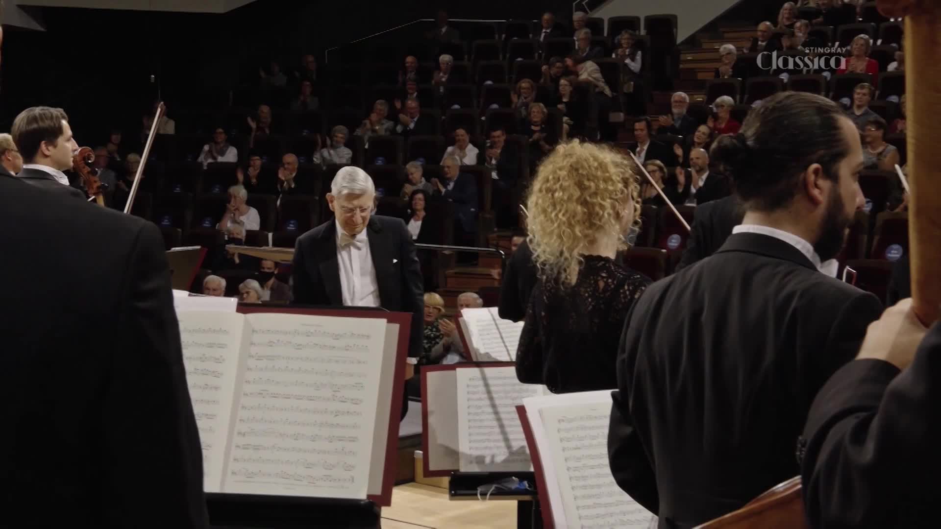 The conductor, a distinguished gentleman in a tuxedo, bows his head as the orchestra prepares to play. Around him, musicians in formal wear adjust their instruments, their sheet music resting on stands. The conductor, a distinguished gentleman in a tuxedo, bows his head as the orchestra prepares to play. Around him, musicians in formal wear adjust their instruments, their sheet music resting on stands.