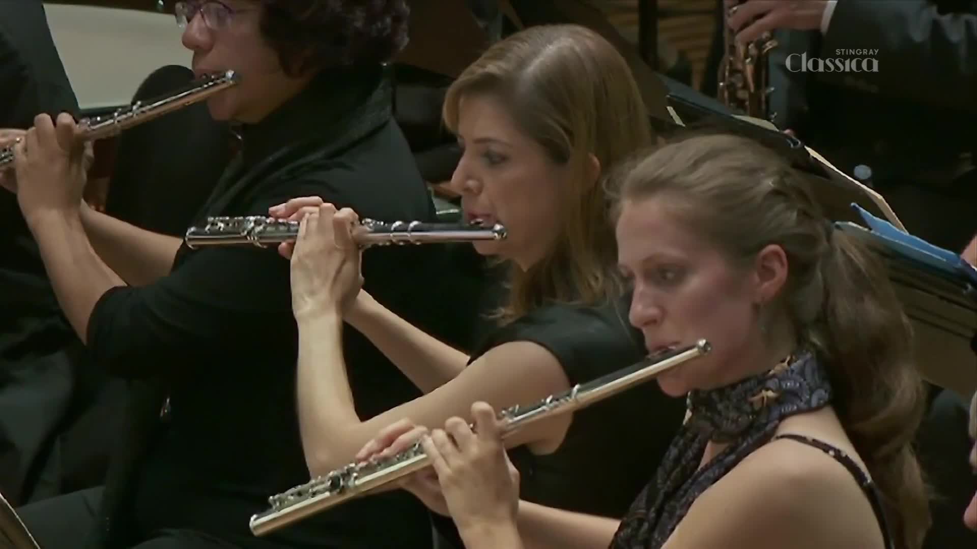 Three women in an orchestra are playing their flutes. The Stingray Classica logo is visible in the background. Three women in an orchestra are playing their flutes. The Stingray Classica logo is visible in the background.