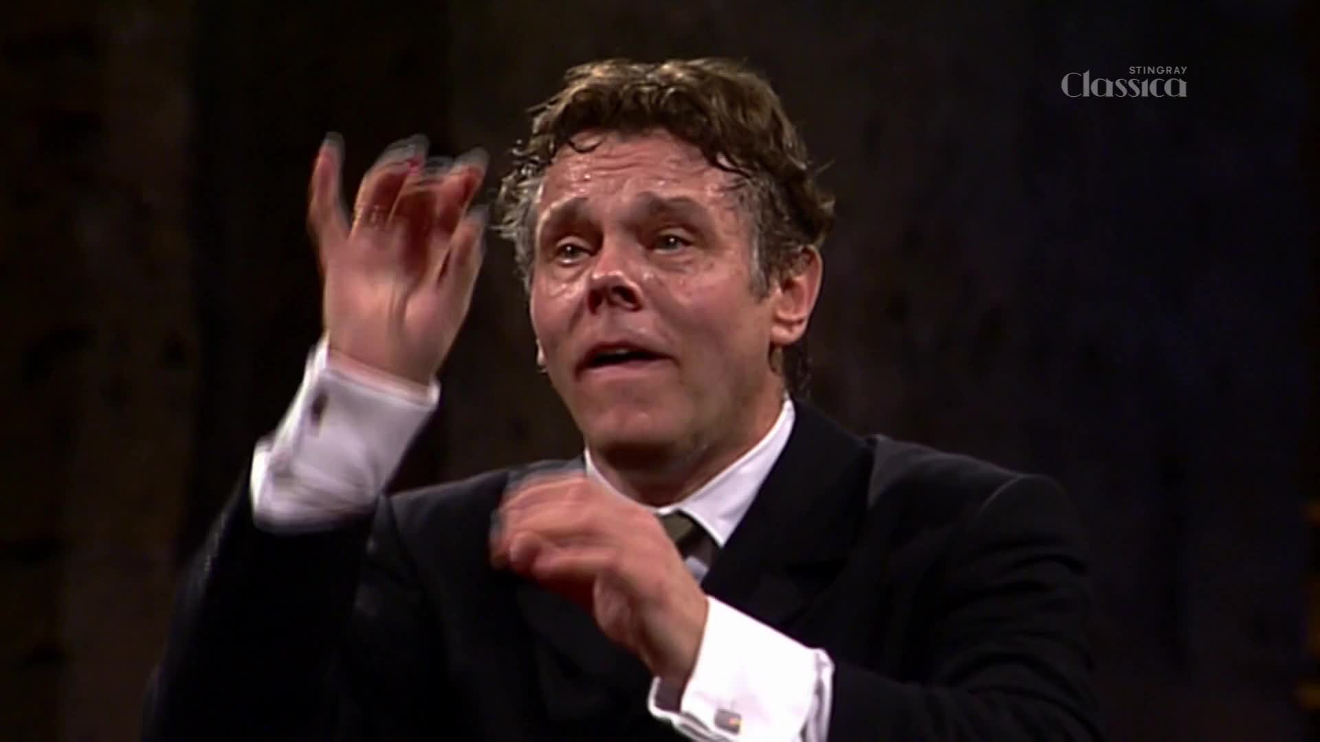 His hands move with urgency, guiding the orchestra through a powerful passage. The Stingray Classica logo appears in the corner, a subtle reminder of the broadcast. His hands move with urgency, guiding the orchestra through a powerful passage. The Stingray Classica logo appears in the corner, a subtle reminder of the broadcast.