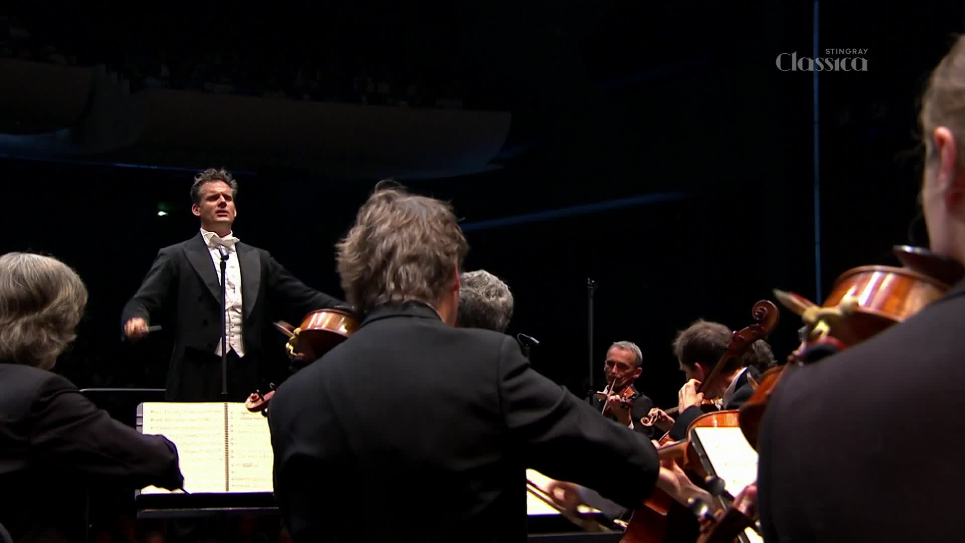 The conductor raises his baton, a crisp white shirt and black tailcoat sharp against the dark stage. Musicians in the foreground lean into their instruments, their bows poised to play.