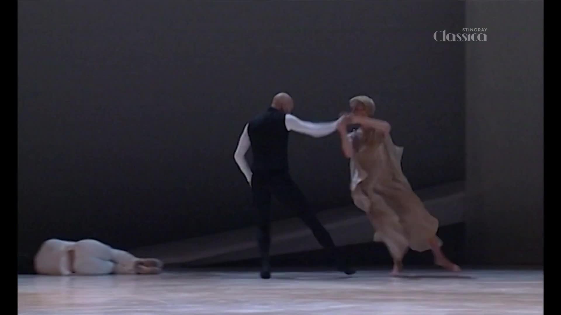 A bald man in a black vest and white shirt grapples with a woman in a flowing tan dress. Another dancer lies still on the floor to the left.