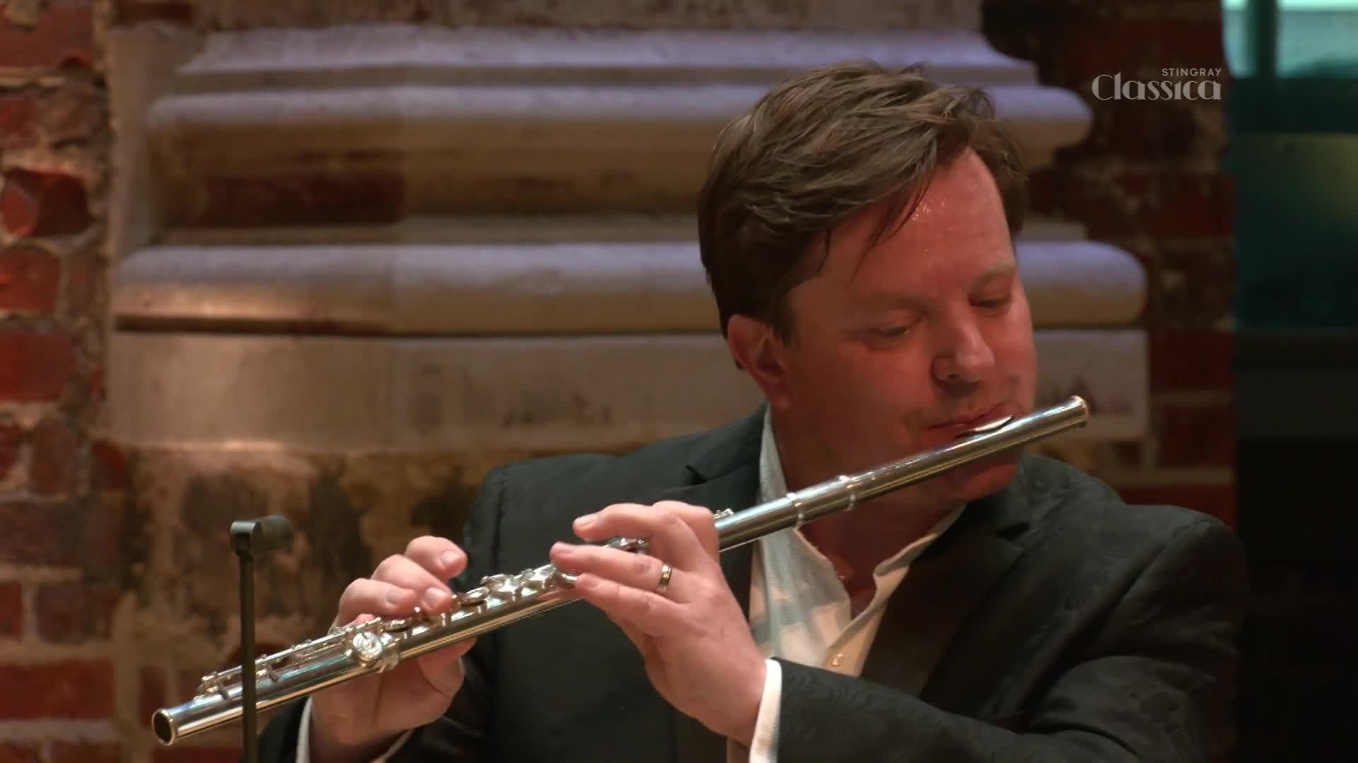 A man in a dark suit plays a silver flute, his fingers moving nimbly across the keys. The Stingray Classica logo is visible in the background, suggesting this performance is for a Canadian audience.