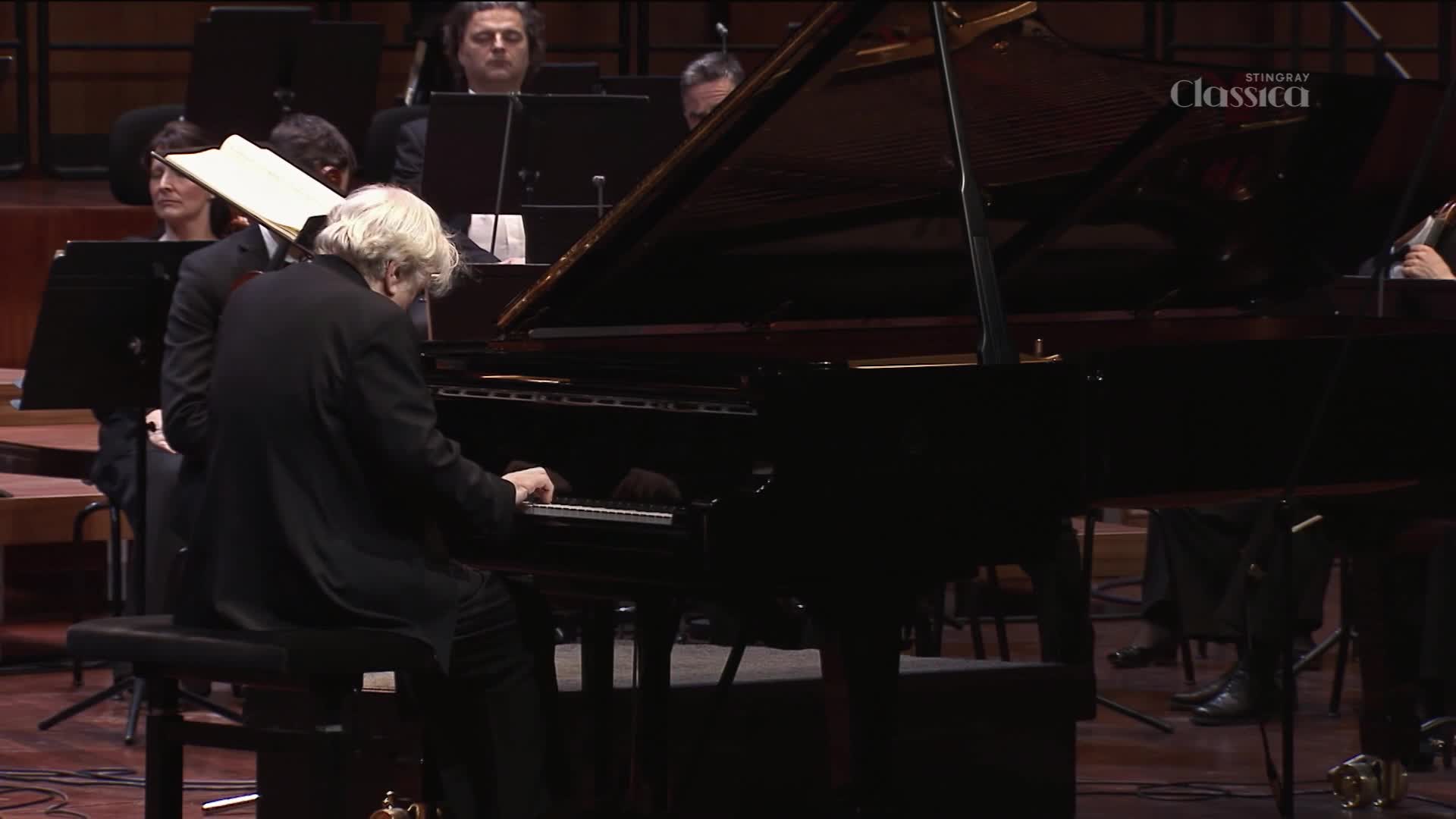 A pianist with flowing white hair plays a grand piano on a stage in Canada. An orchestra sits behind him, ready to accompany the performance.