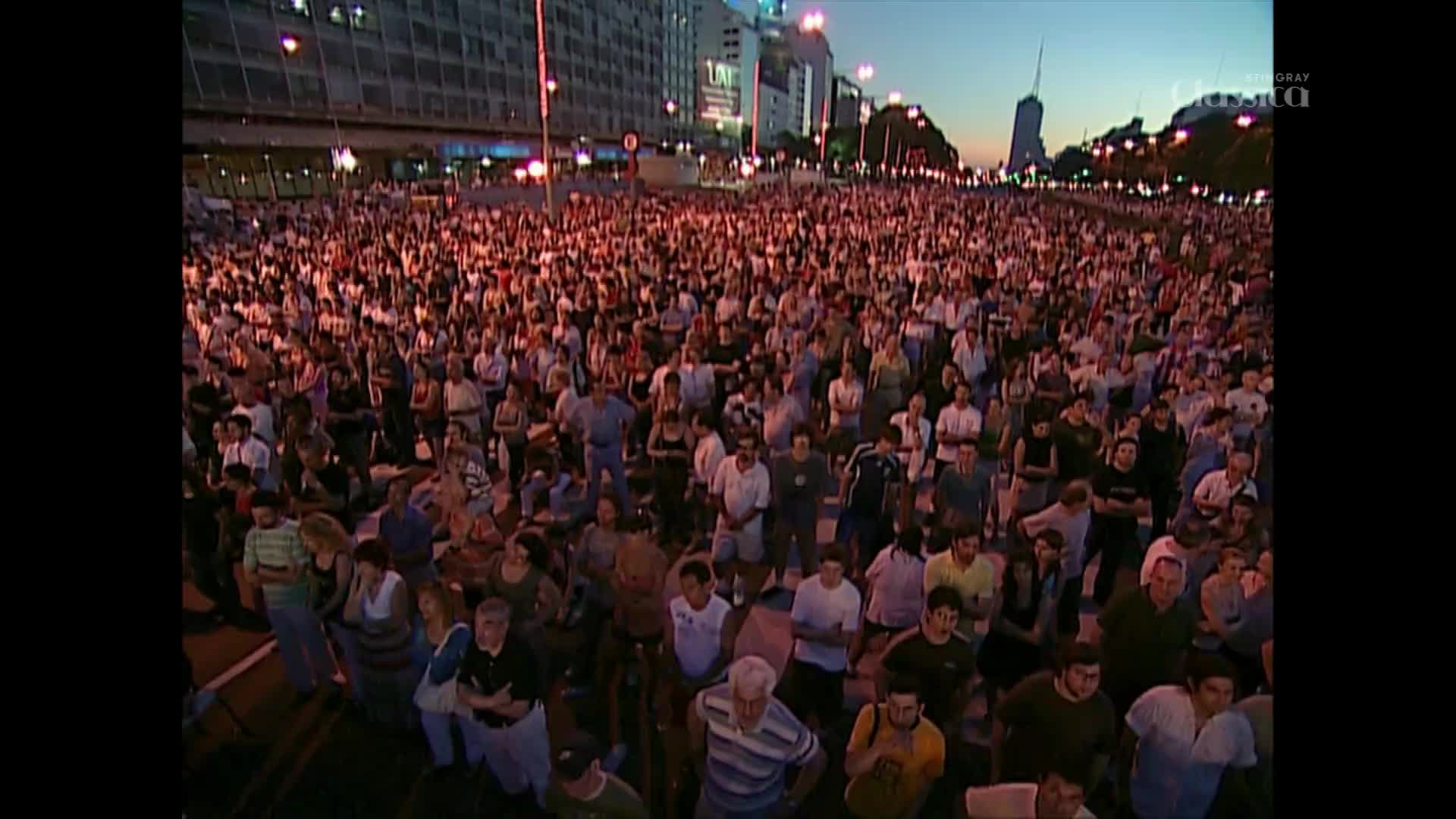 A massive crowd fills the plaza, their faces illuminated by the warm glow of streetlights. The sky above is a soft twilight blue, hinting at the end of a summer day in Canada.