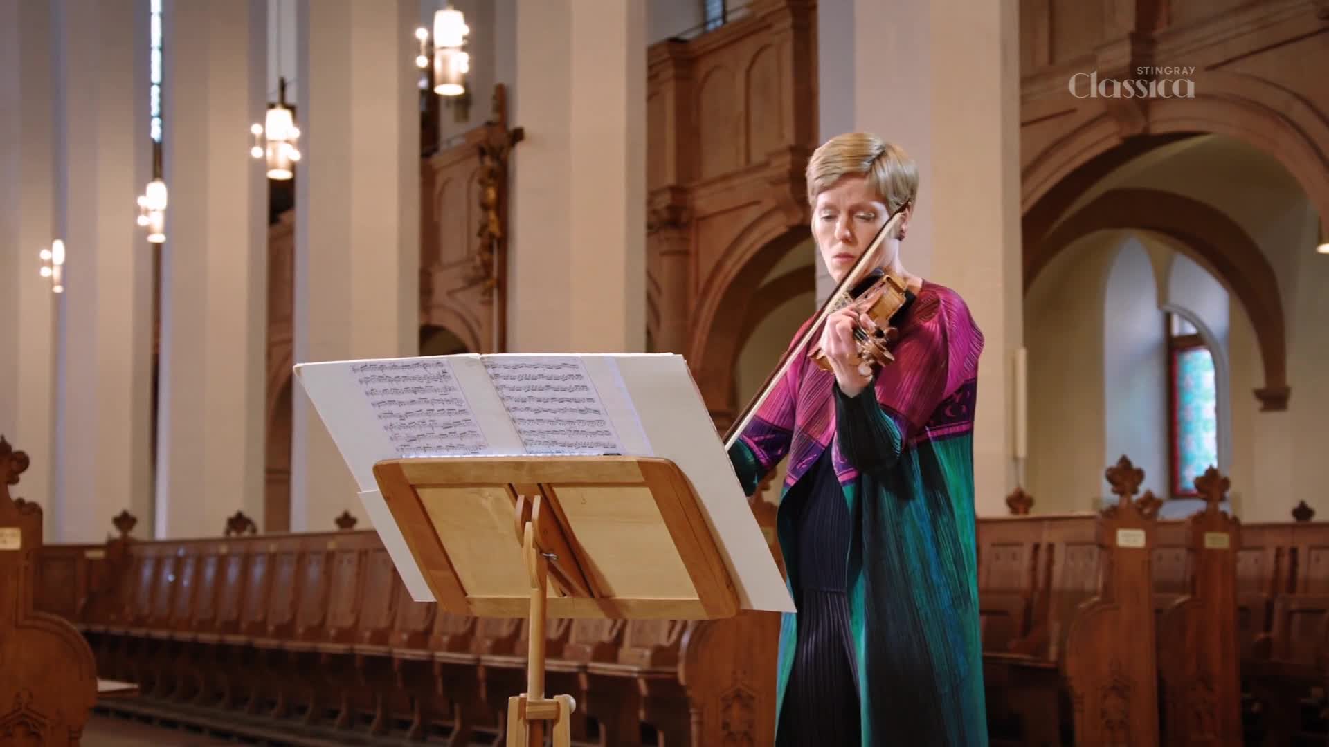 A woman plays the violin in a church, her bow moving across the strings. The Stingray Classica logo is visible in the upper right corner.