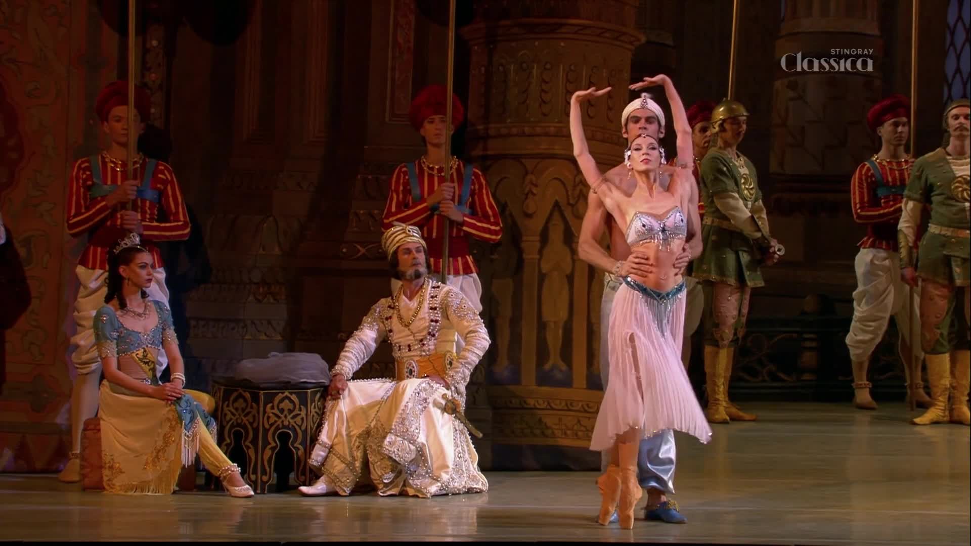 A dancer in a pale pink skirt pirouettes, supported by her partner. Beside them, a man in ornate white robes sits regally, while others in colorful uniforms stand guard.