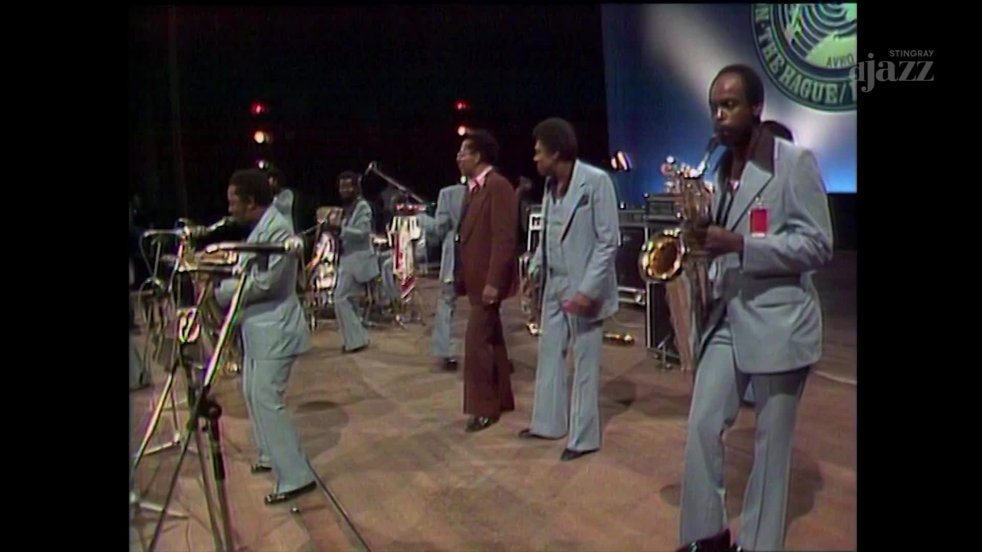 A band in matching light blue suits is on stage, some with instruments raised. The saxophonist is blowing into his horn, the sound likely echoing across the concert hall.
