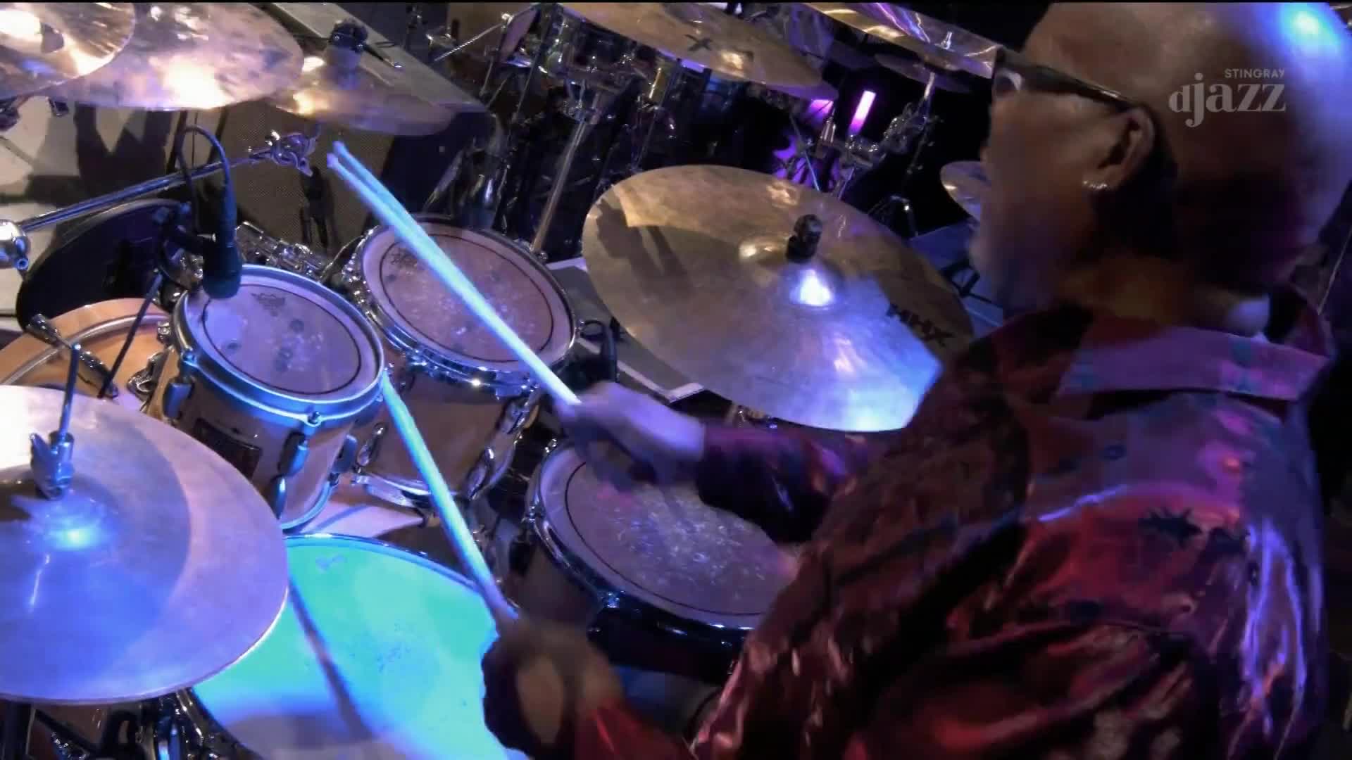 The drummer's sticks are a blur as they strike the snare and toms. The Stingray DJAZZ logo is just visible in the corner, and the stage lights are reflecting off the cymbals.
