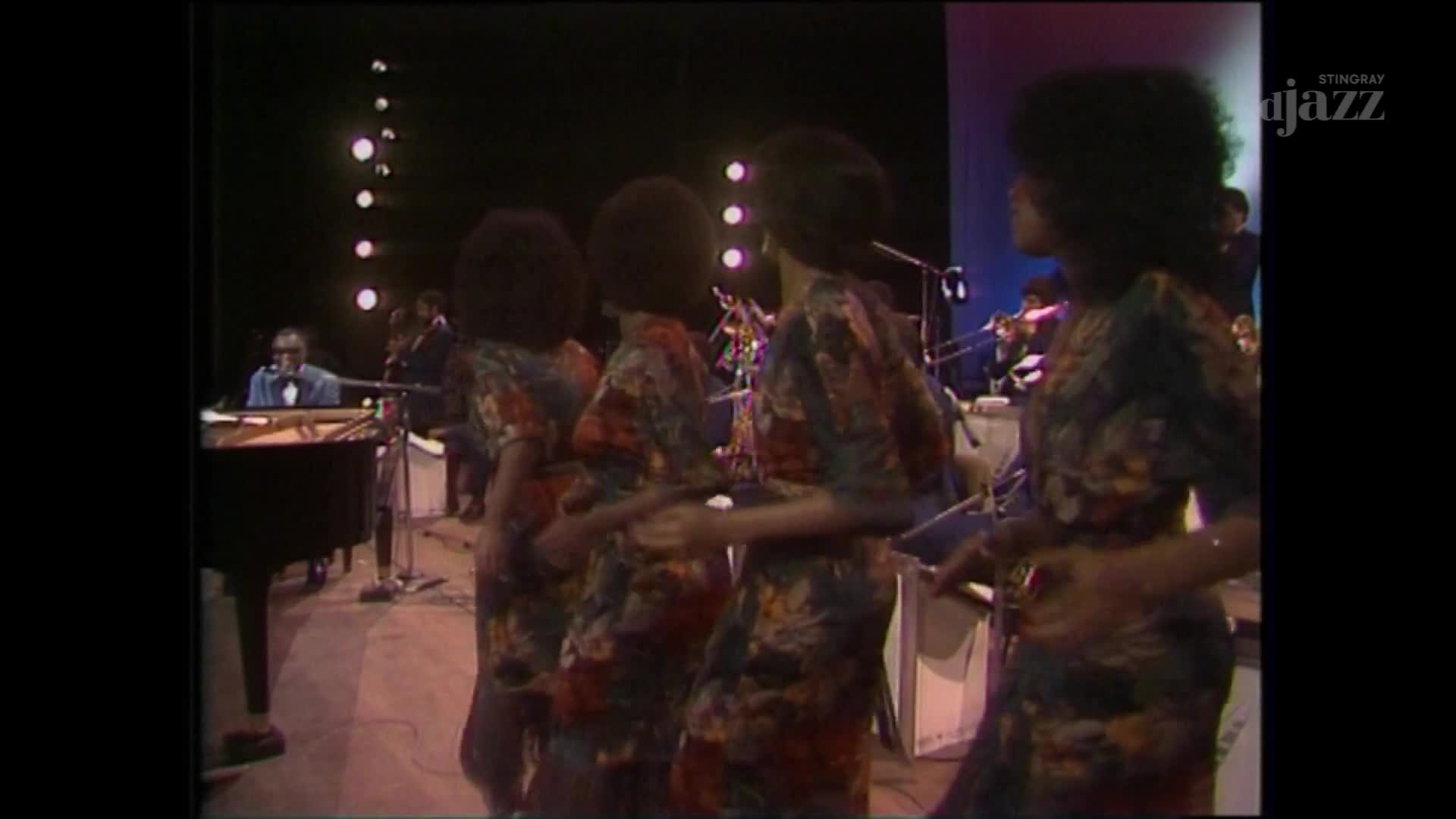 The piano player, in a blue suit, is focused on the keys. Four women in patterned dresses sway in unison, their backs to the camera.

