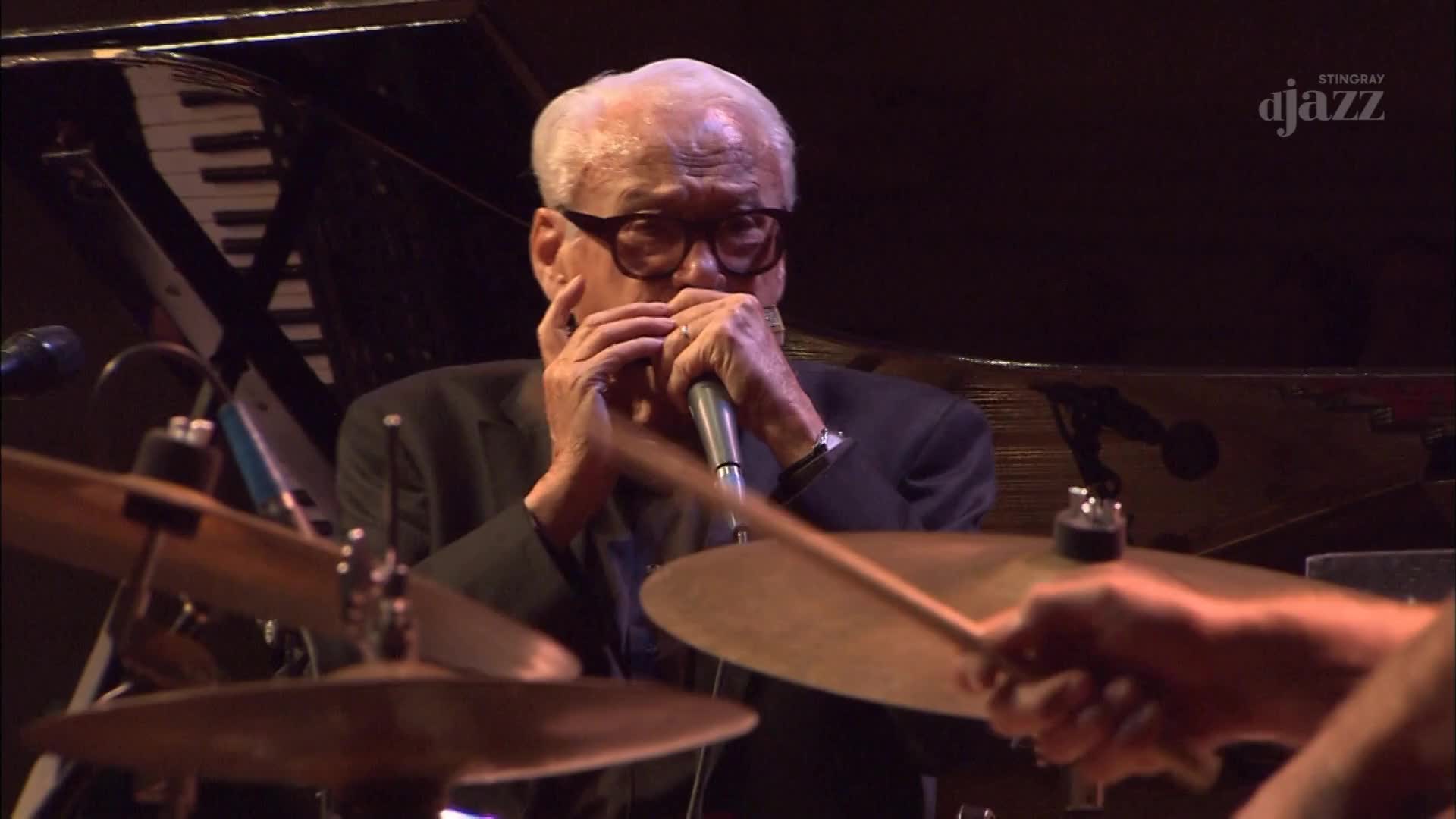 The harmonica player, framed by his glasses, presses the instrument to his lips. A drummer's stick arcs down, poised to strike the cymbal.
