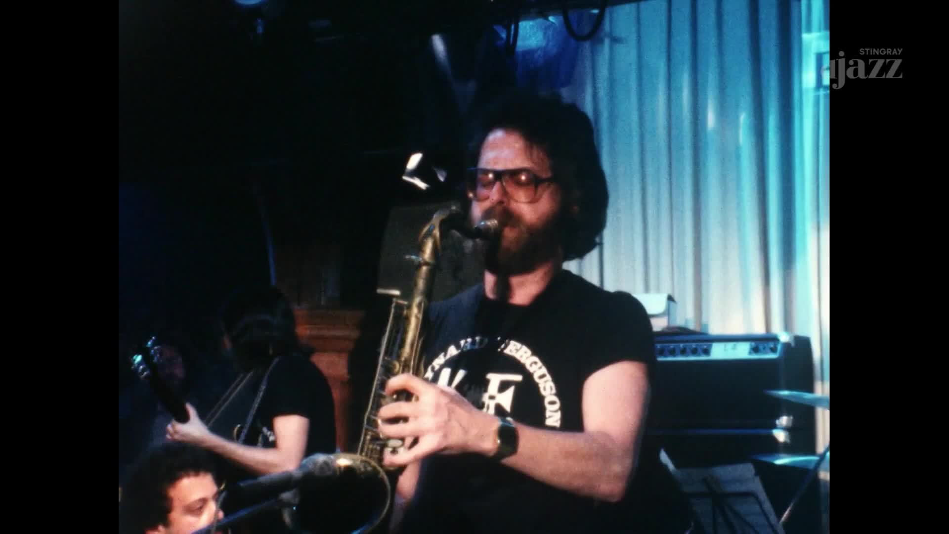 A man with a beard and glasses blows into a saxophone, his fingers dancing over the keys. The band plays, bathed in the blue light of the stage.
