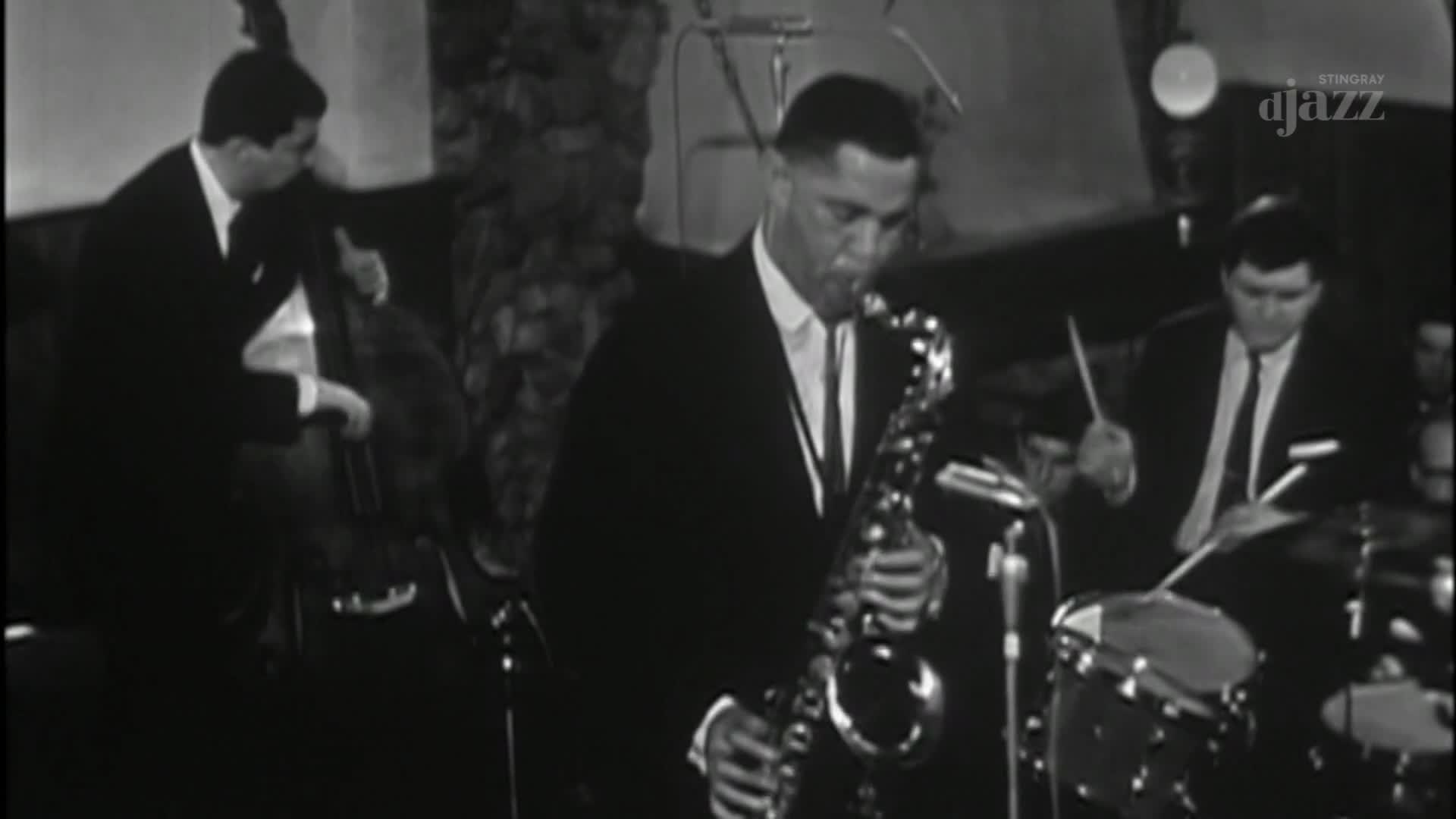 The saxophonist, in a dark suit, blows into his instrument, his fingers dancing over the keys. A bassist and drummer, also in suits, accompany him, their instruments filling the small venue with music.
