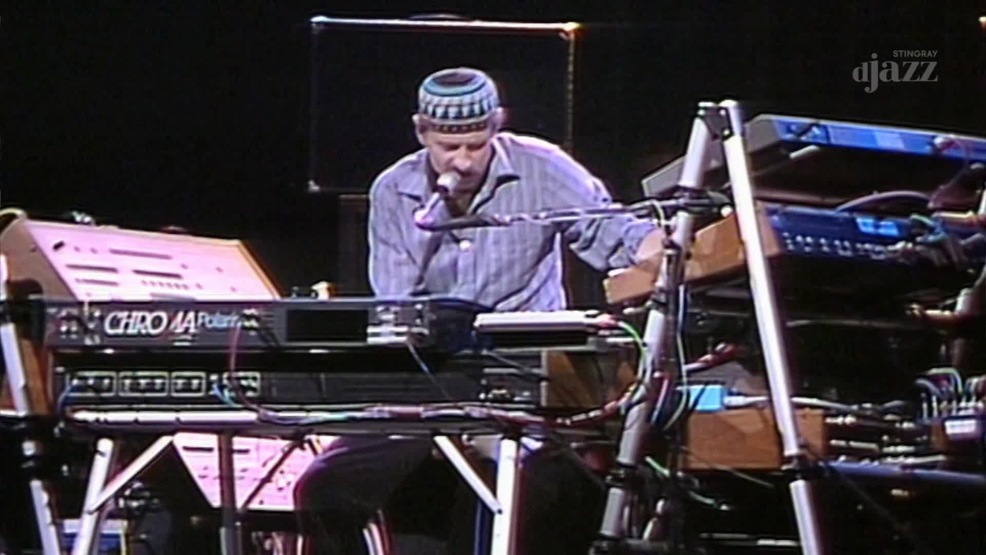 A musician in a patterned cap leans into a microphone, his fingers dancing across the keys of a Chroma Polaris synthesizer. The Stingray DJAZZ logo is visible, and the stage is set for a performance.
