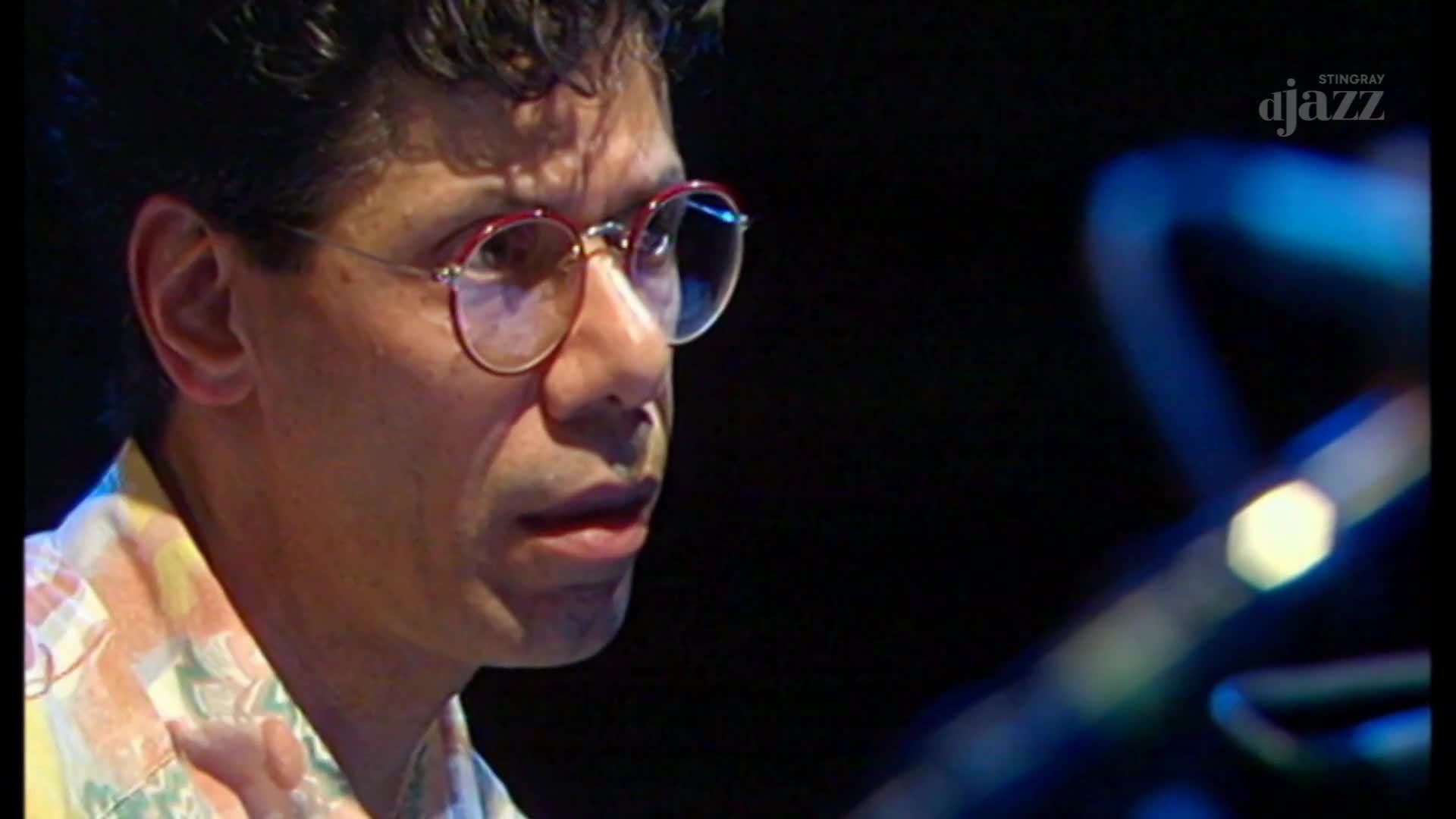 The musician's face is lit by a stage light, his glasses reflecting the blue glow. He's focused, eyes fixed on something just out of frame, and the logo for Stingray DJAZZ is visible in the corner.
