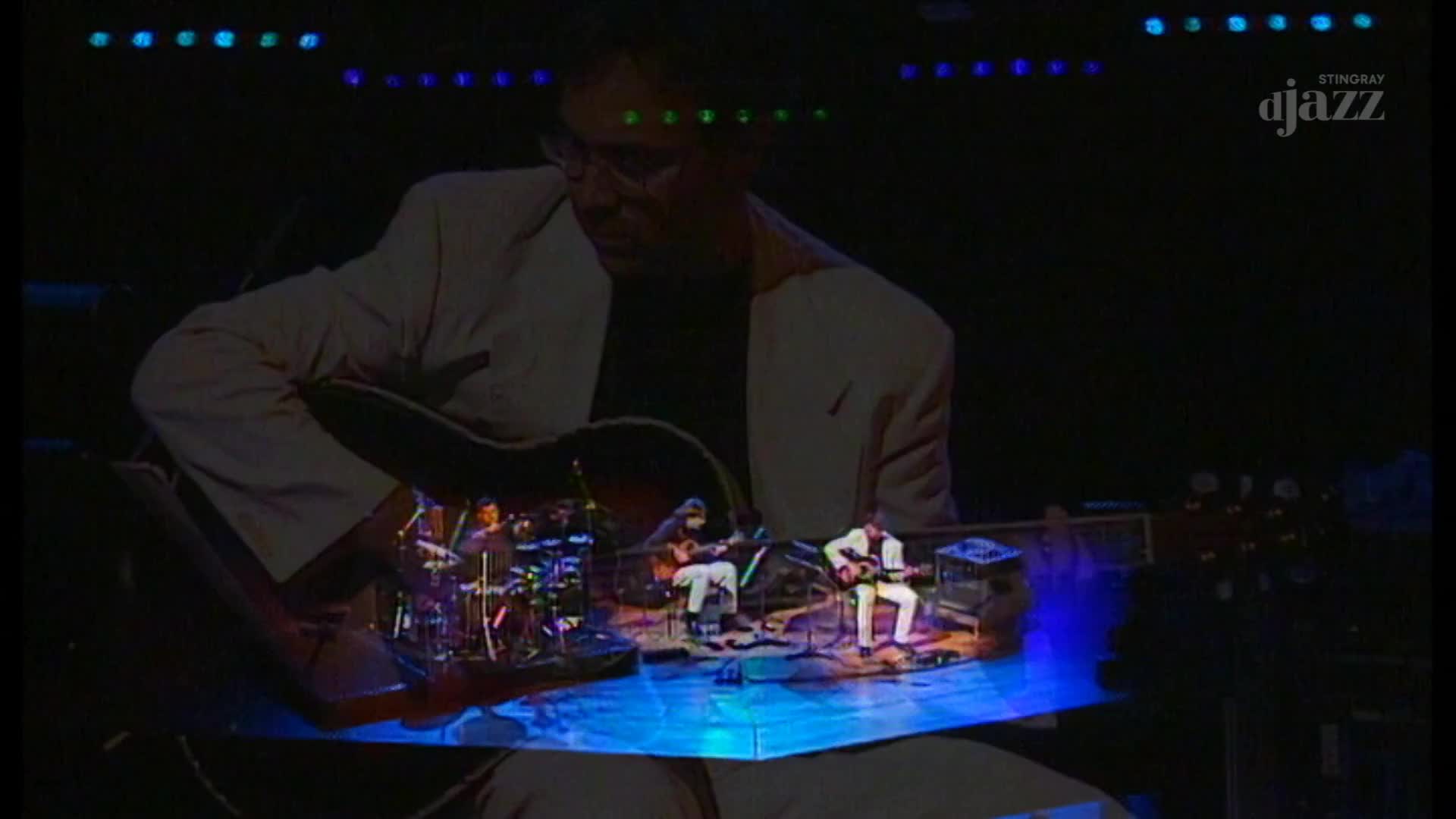 The guitarist in the white jacket strums his instrument, illuminated by the stage lights. A smaller stage scene is reflected in the guitar, where a band plays on a blue-lit platform.
