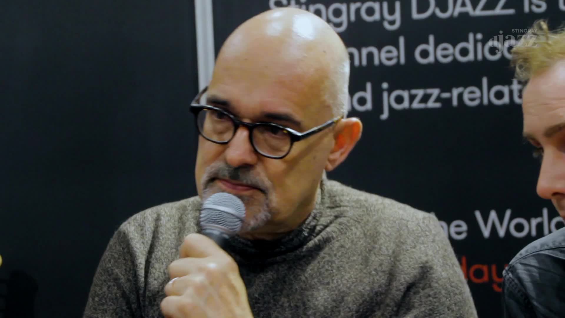 The man with the glasses holds a microphone, speaking, while the Stingray DJAZZ logo is visible behind him. Beside him, another person listens intently.

