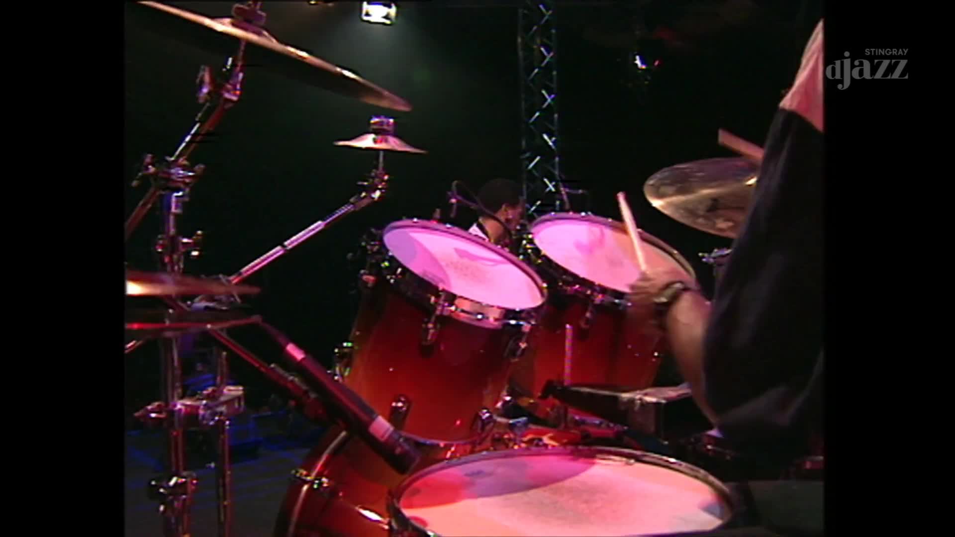 The drummer's arms are a blur as sticks strike the drums, the red of the shells reflecting the stage lights. A Stingray DJAZZ logo is visible in the corner, a reminder of where this Canadian jazz performance is being broadcast.
