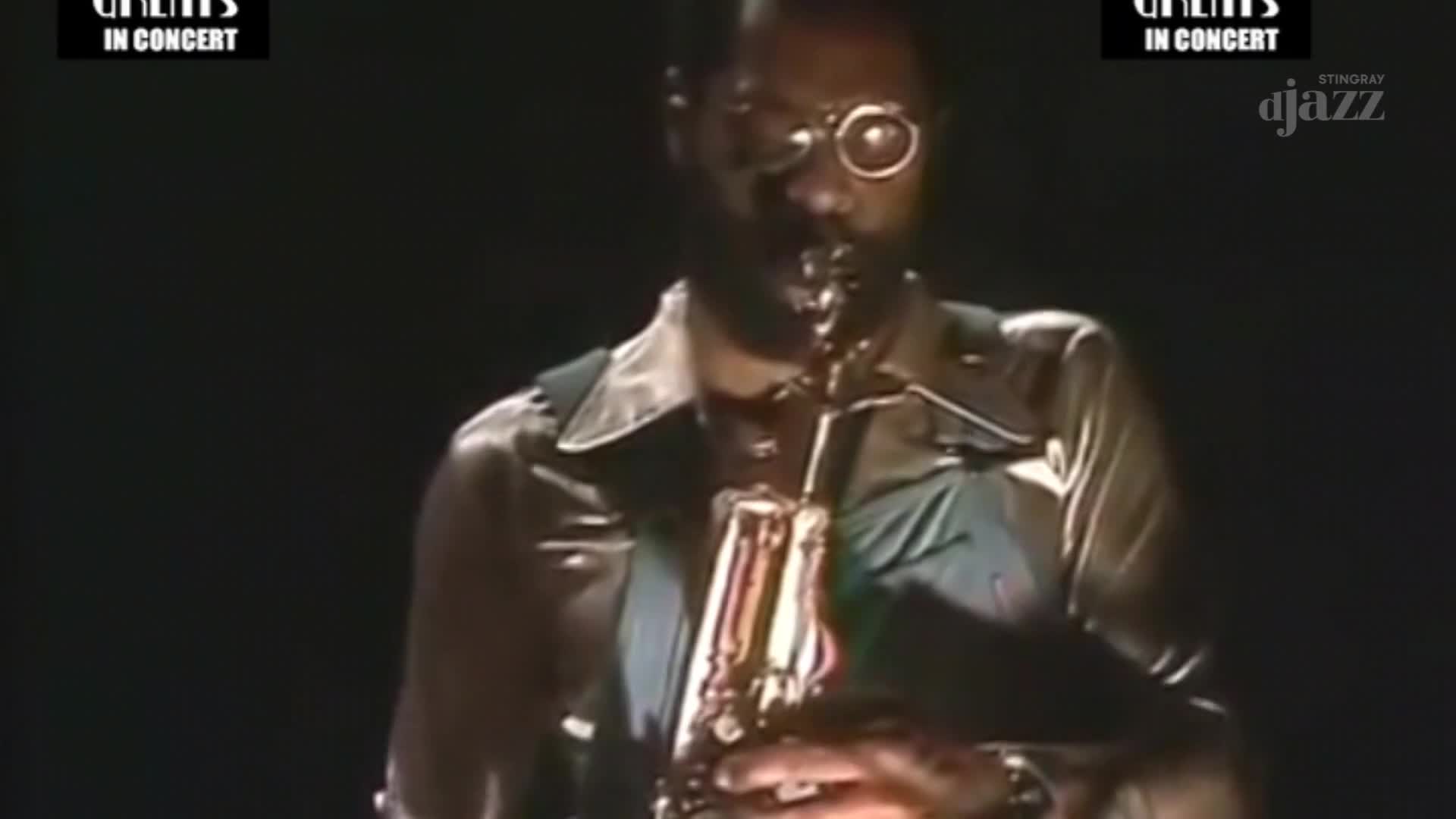 The musician, wearing a single monocle, holds a saxophone, his fingers moving over the keys. The Stingray DJAZZ logo is visible, and the concert is likely from Canada.
