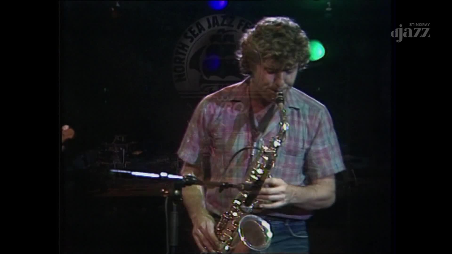 A man in a plaid shirt holds a saxophone, his breath visible as he plays. The Stingray DJAZZ logo is visible, and the North Sea Jazz Festival banner hangs in the background.
