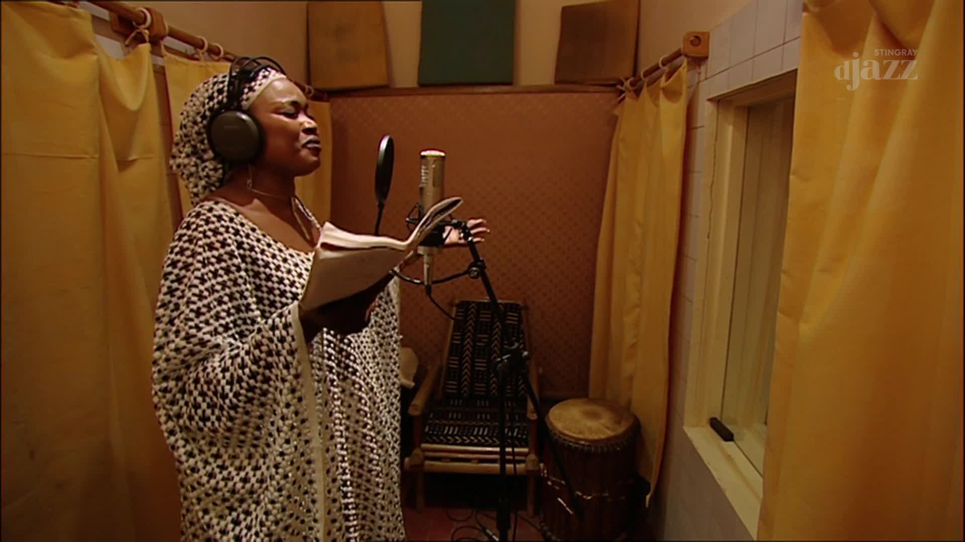 In a dimly lit studio, a woman in a patterned dress stands before a microphone, singing. She's wearing headphones, holding a script, and her voice fills the room.
