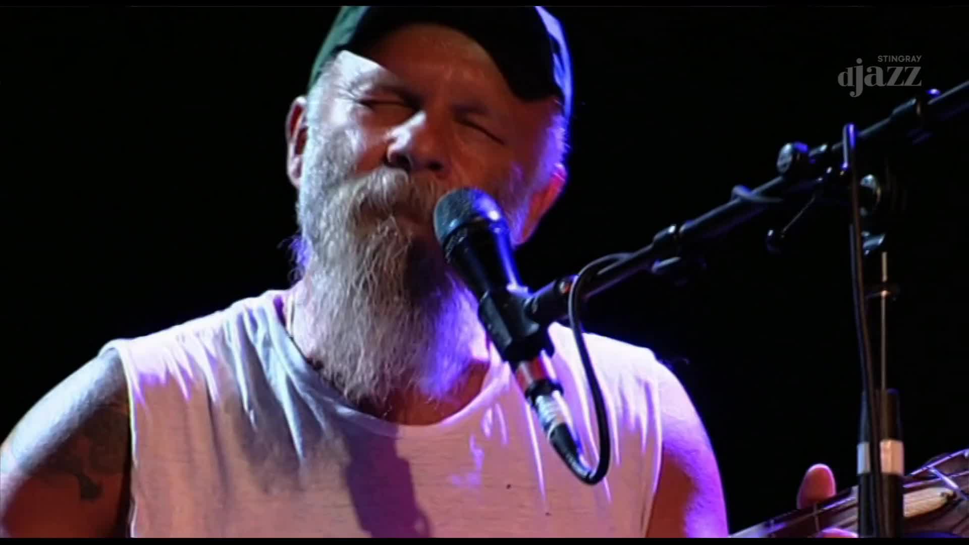 The musician, in a white tank top and cap, is singing, his eyes closed, into a microphone. His fingers pluck at the strings of an instrument, likely a guitar, as the music swells.
