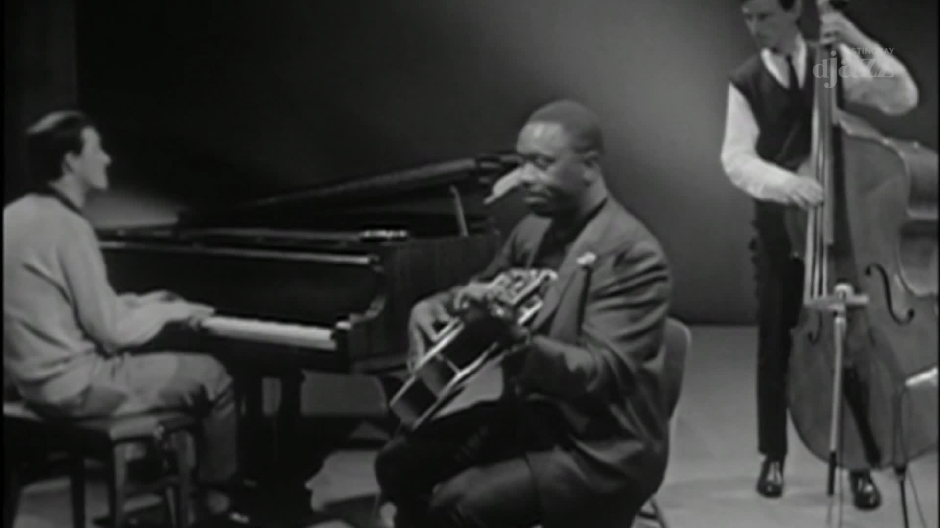 A man in a suit strums a guitar as another plays the upright bass. The pianist's fingers dance across the keys, the music filling the studio.
