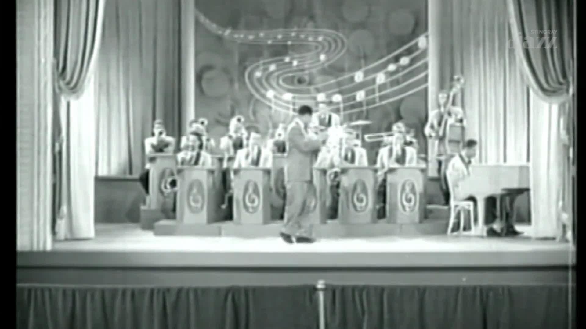 A band in suits plays on a stage, the musicians behind decorative podiums. The man in the center, holding a horn, takes a step forward as the music swells.
