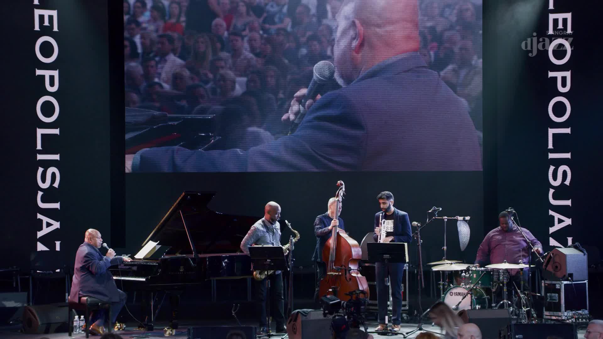 The band is mid-performance, with a large screen behind them showing a close-up of a singer. The musicians are lit by stage lights, their instruments gleaming as they play.
