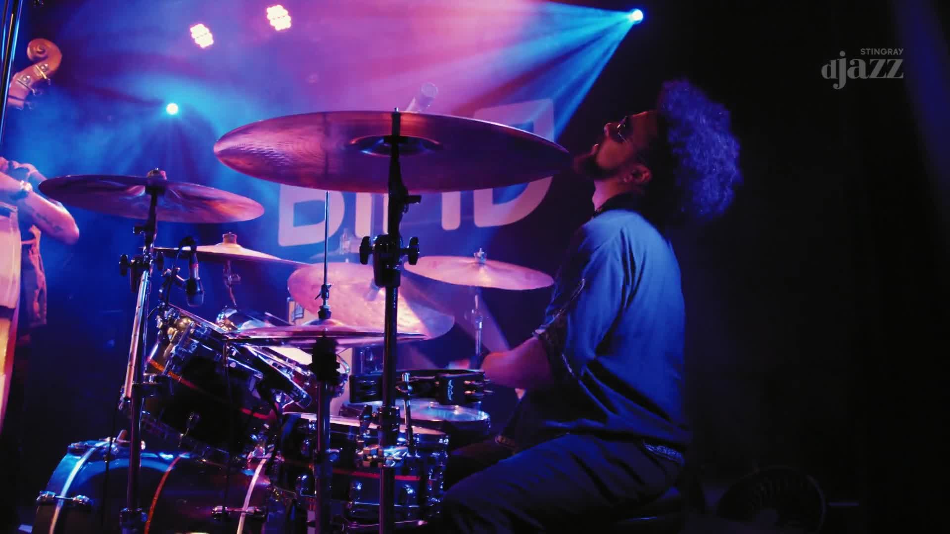 The drummer, bathed in purple and blue light, is mid-swing, his sticks striking the cymbals. A Stingray DJAZZ banner hangs in the background, as the music fills the room.
