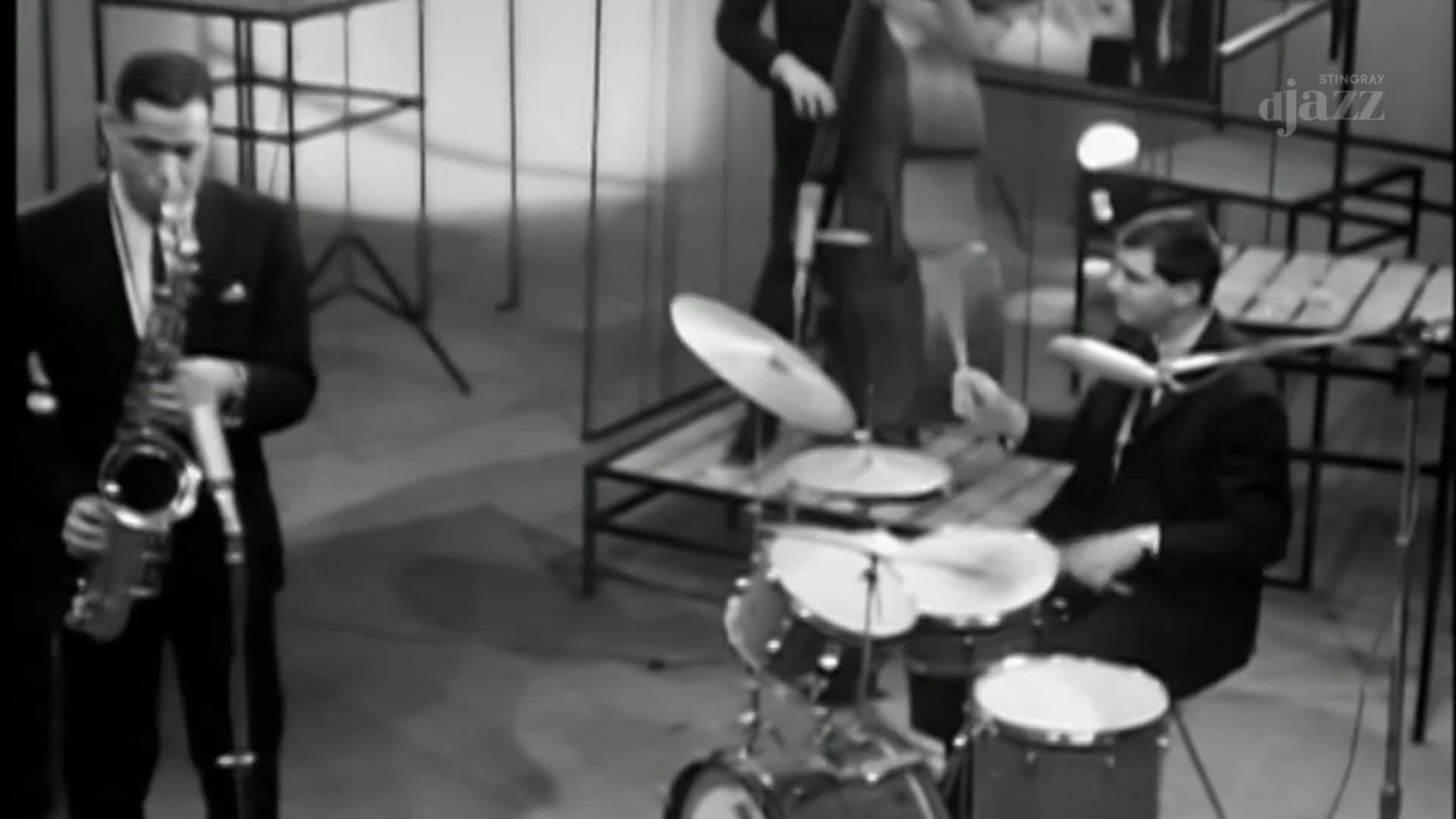 The drummer's sticks are a blur as he hits the cymbals and drums. A saxophone player, in a dark suit, is poised to play, microphone at the ready.
