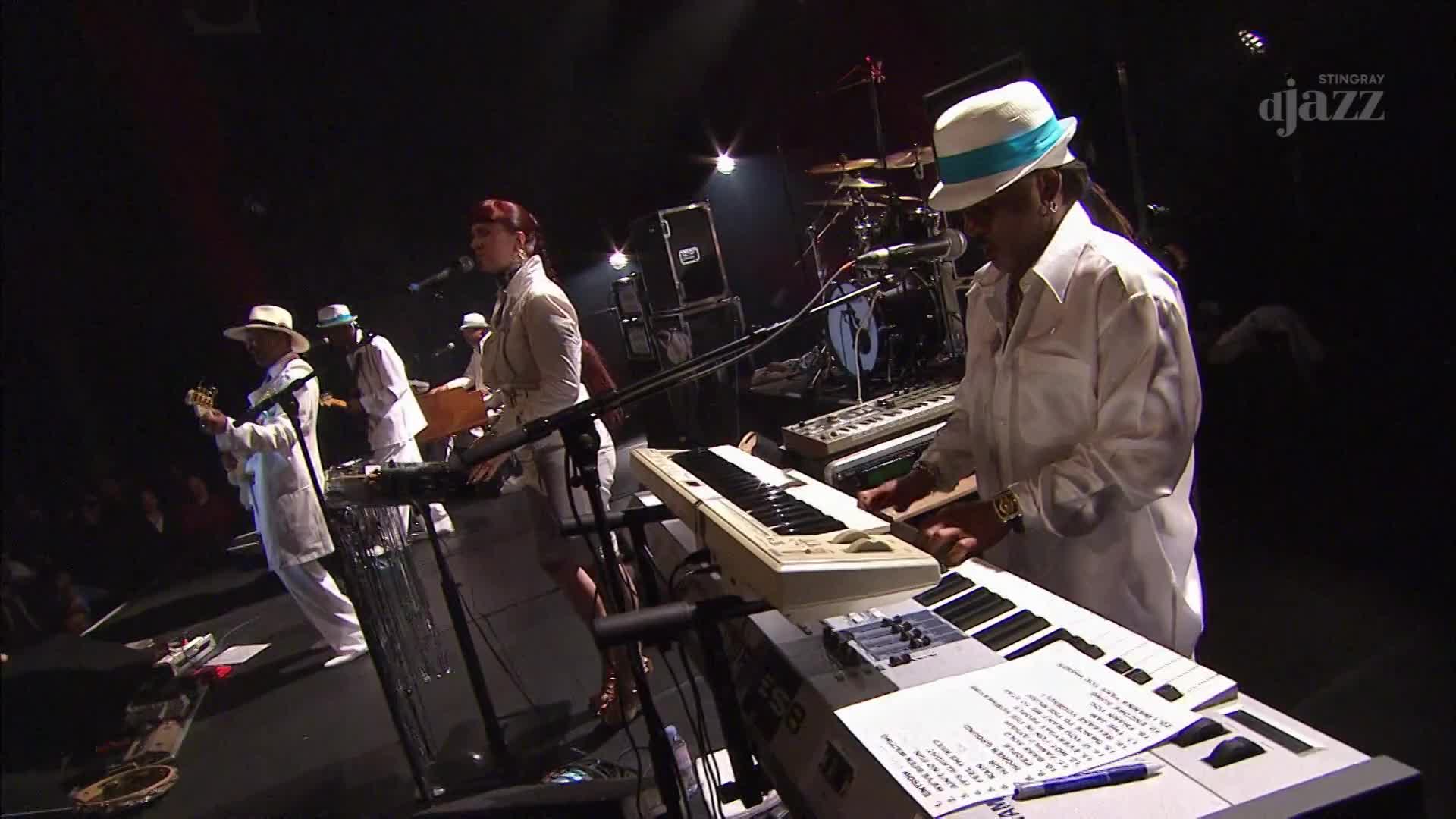 A band in white suits and hats is playing on a stage, under the Stingray DJAZZ logo. The keyboardist in the foreground is focused, his fingers dancing over the keys.
