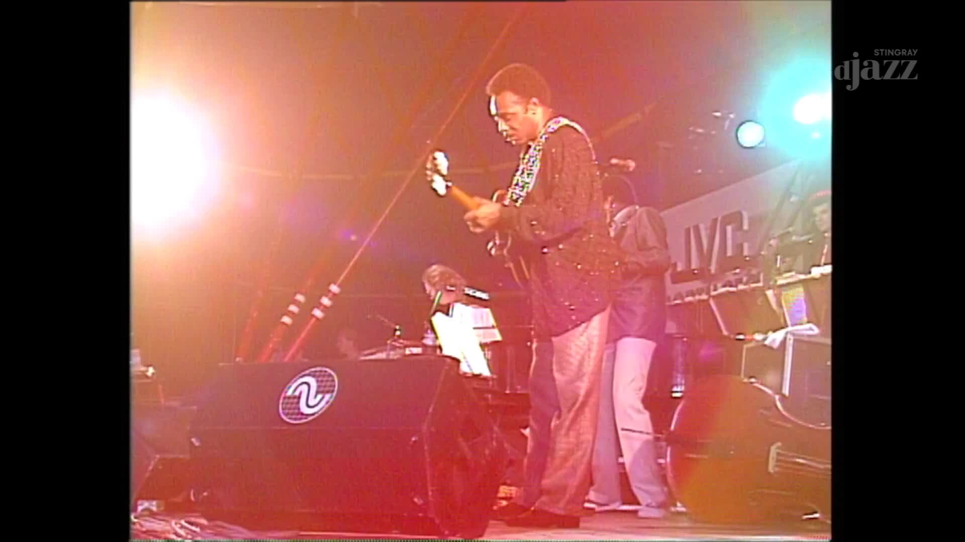 A musician in a patterned shirt strums a guitar, bathed in stage lights. Behind him, another player stands near a double bass, all part of the performance broadcast by Stingray DJAZZ in Canada.
