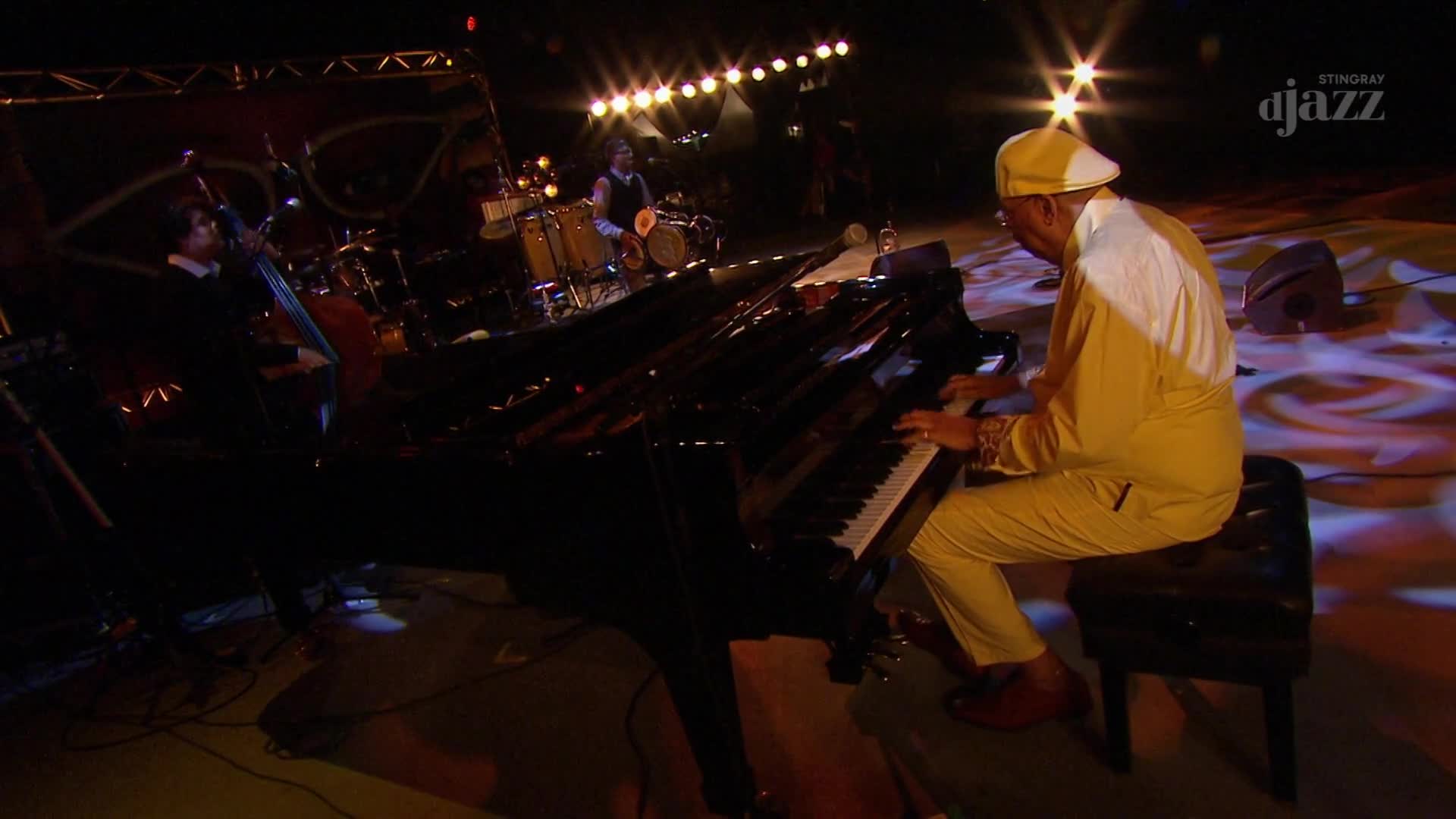 The pianist, in a yellow suit and cap, is bent over the keys of a grand piano. A bassist and drummer are in the background, illuminated by stage lights, as part of a performance for Stingray DJAZZ in Canada.
