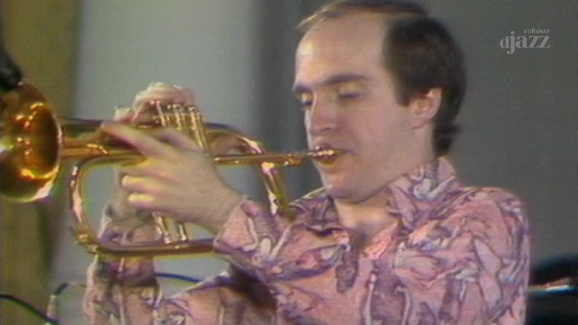 A man in a patterned shirt holds a trumpet, his lips pressed against the mouthpiece. He's playing, the instrument glinting under the lights, a scene captured by Stingray DJAZZ in Canada.
