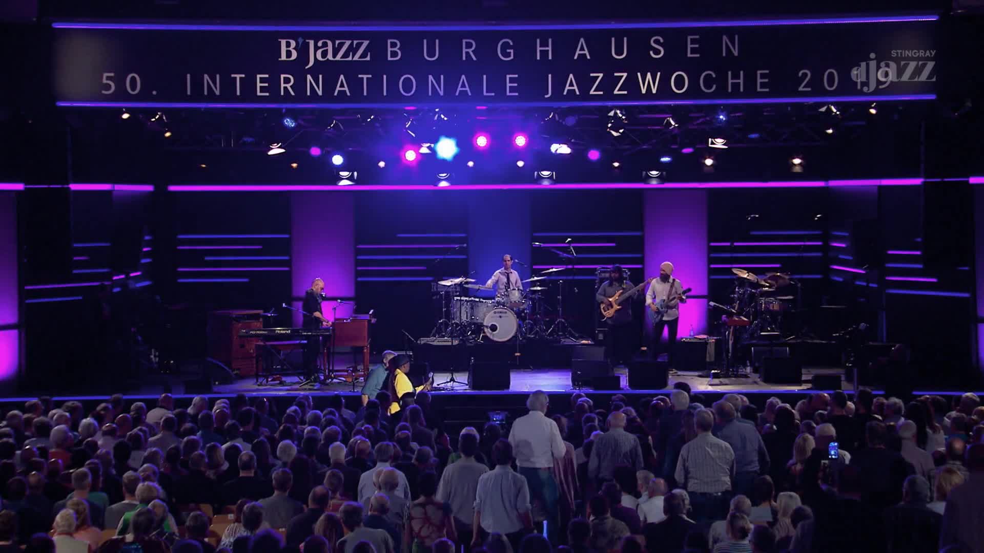 A jazz band is performing on stage, bathed in purple and blue light, for the 50th International Jazz Week. The crowd in front of me is packed, many with phones up, capturing the music.
