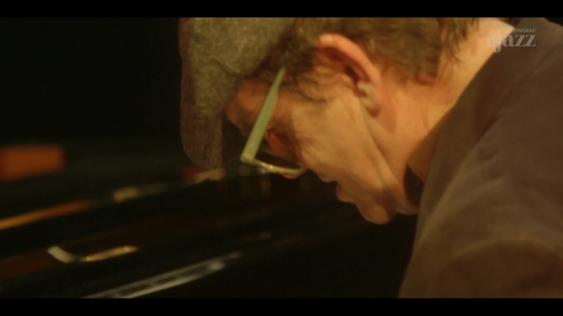 A man with glasses and a cap leans over a piano, his face illuminated by warm light. His fingers, barely visible, seem poised above the keys.
